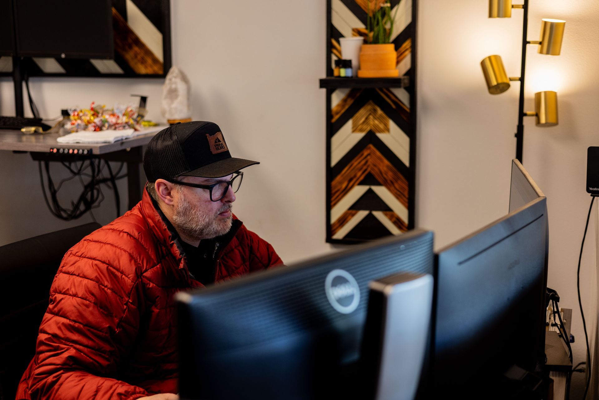 Man in red jacket, glasses, and hat works at computer with dual monitors in office setting.