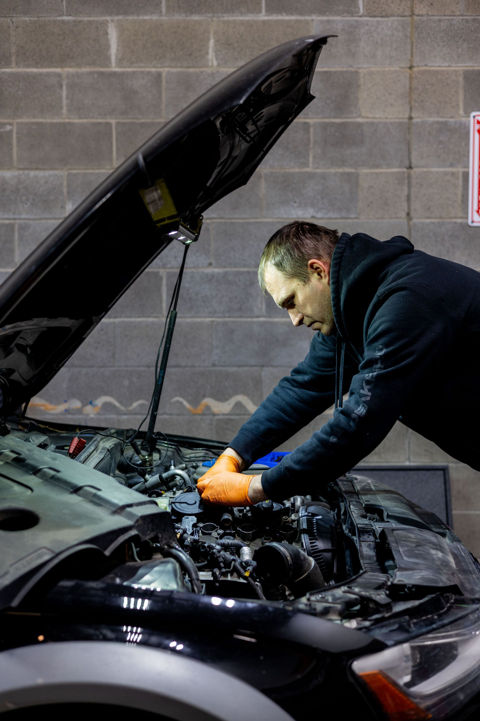 Mechanic with orange gloves working on a black car engine in a garage.| Peak Euro
