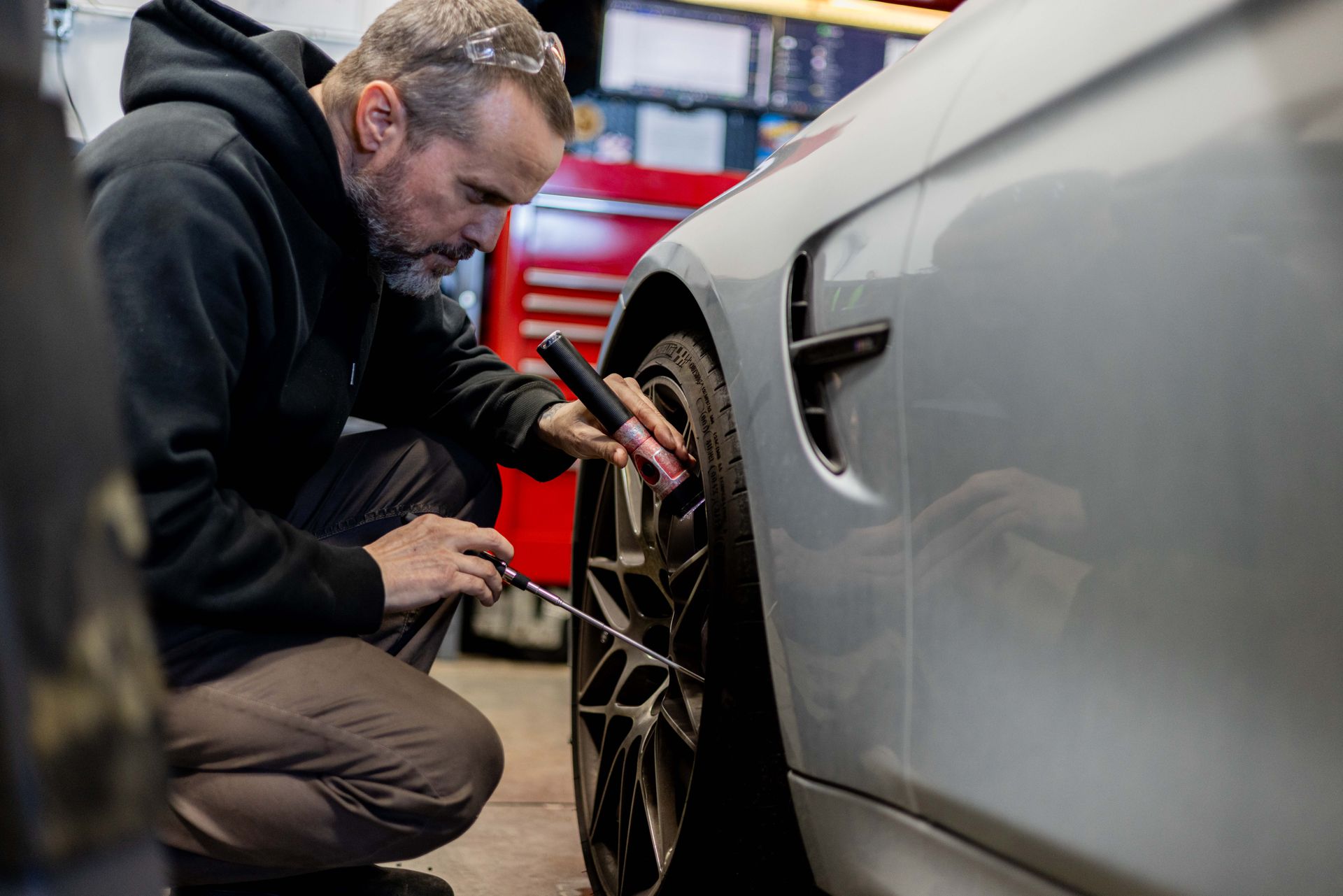 Mechanic in black hoodie checks tire pressure on a silver car in a garage.| Peak Euro