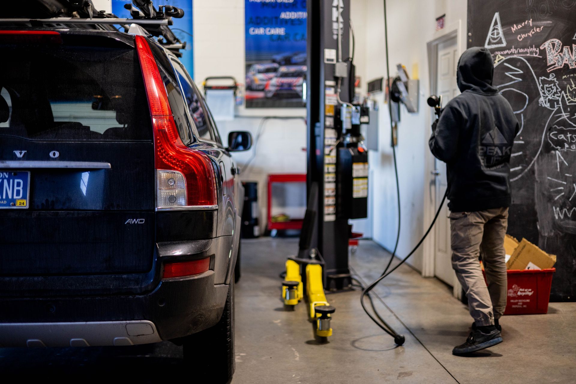 A person in a hoodie holds a hose in a car repair shop next to a Volvo SUV.| Peak Euro