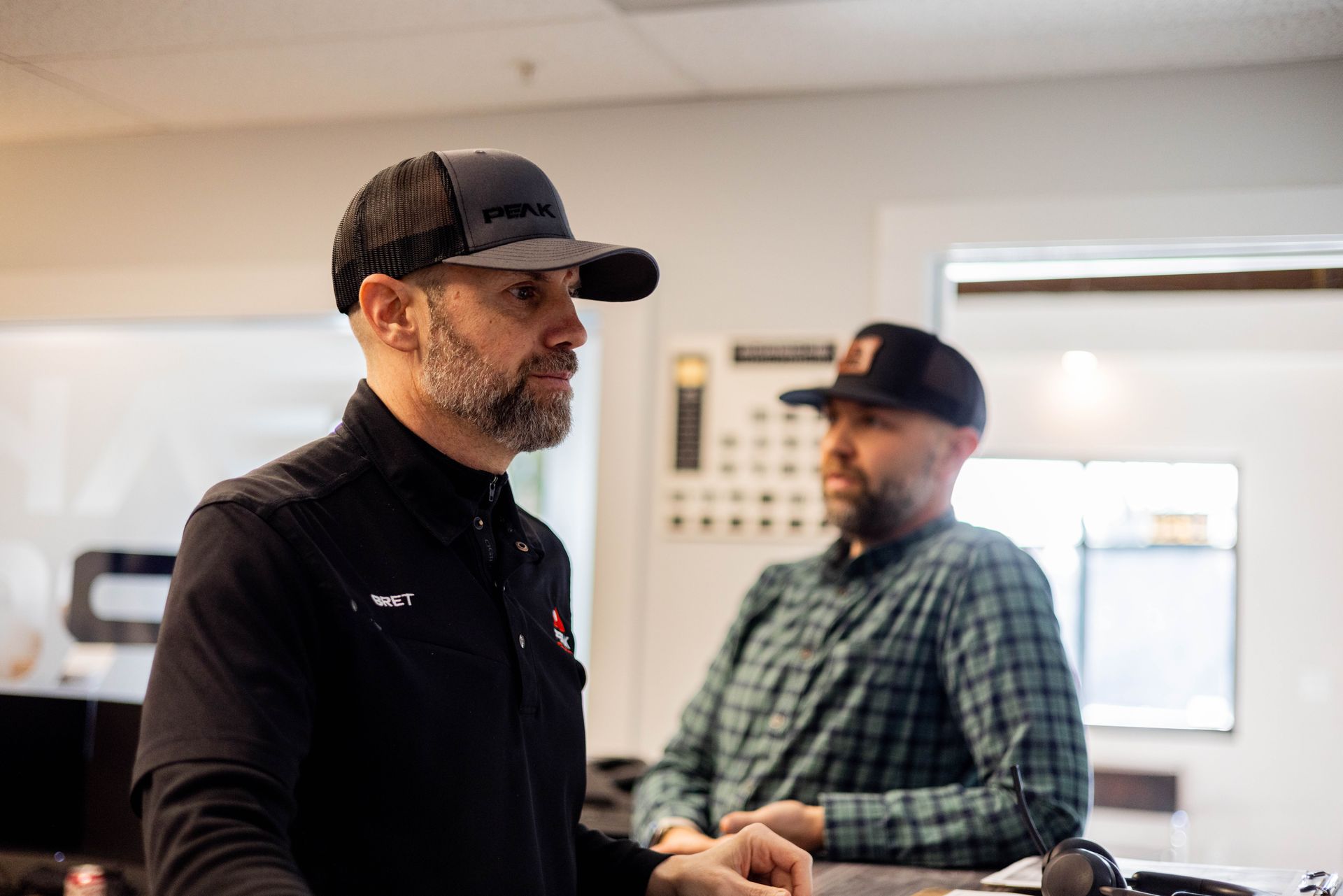 Man in black hat and jacket in a room, looking left. Another man in a hat and plaid shirt in the background.