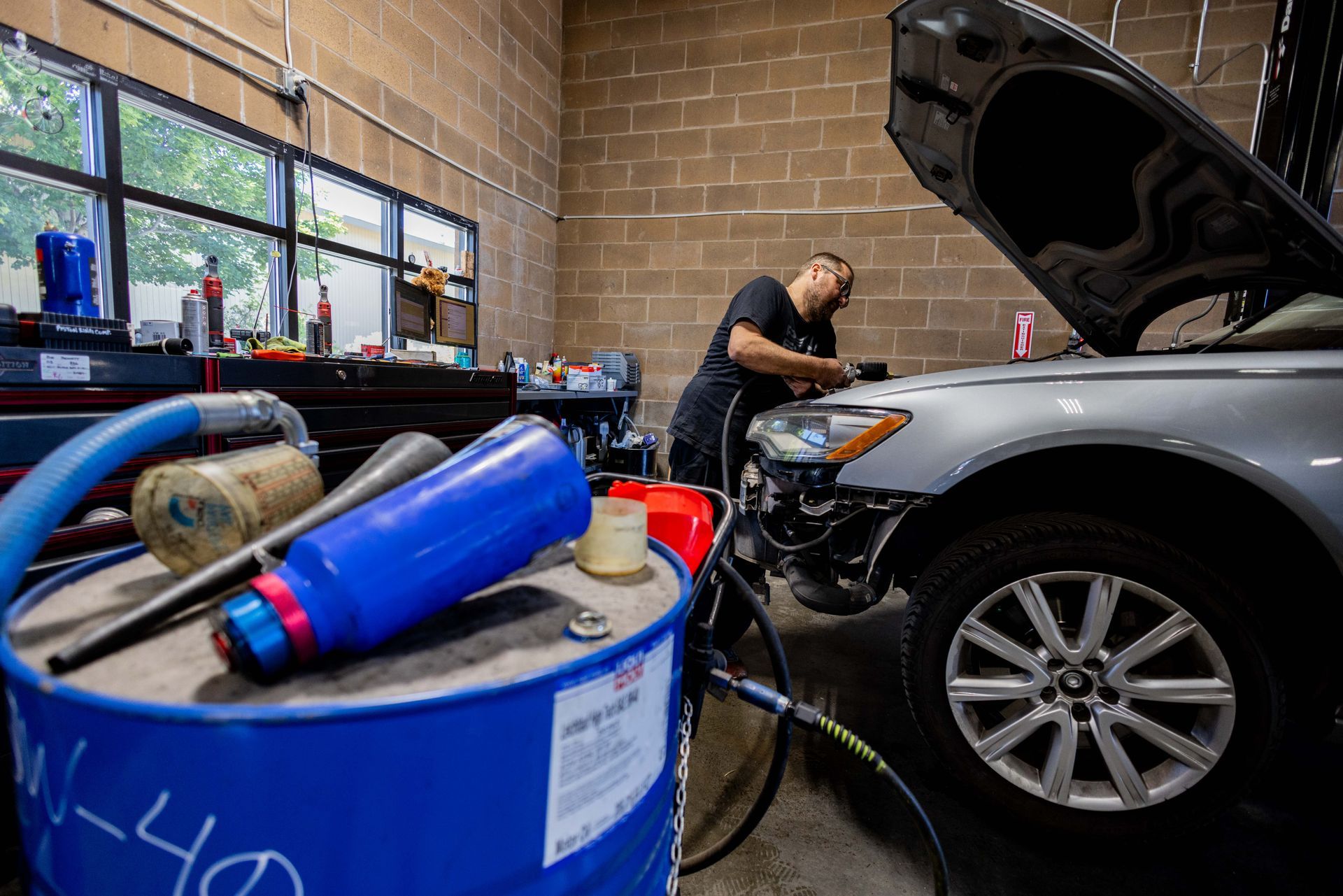 Mechanic working on a silver car in a garage. Blue barrel with tools in the foreground. | Peak Euro