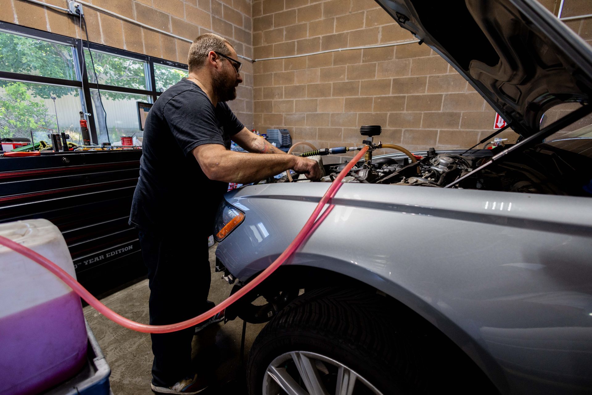 Mechanic working on a silver car with the hood up, using a pink hose. Brick wall background. | Peak Euro