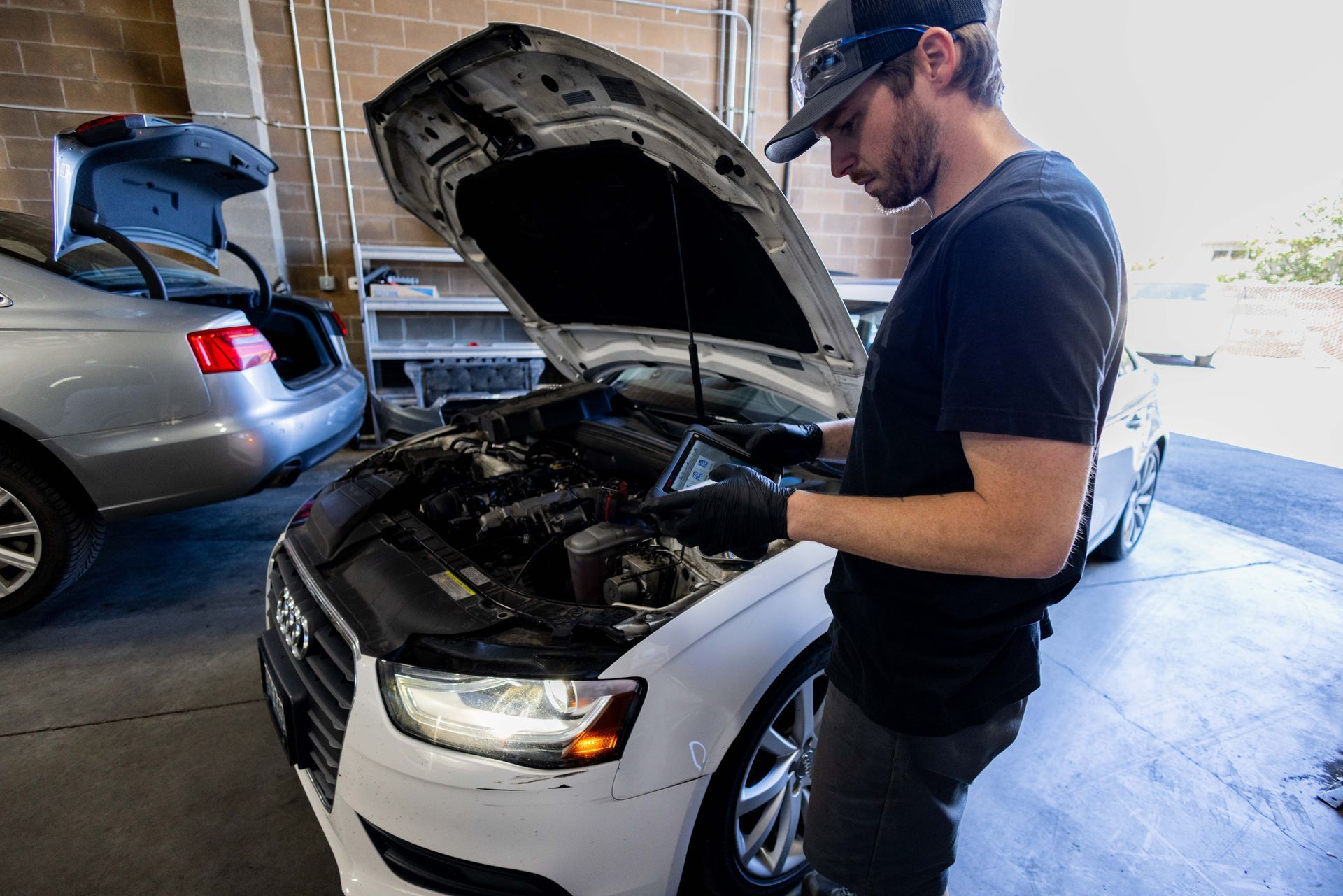Mechanic working on a white car with the hood open in a garage, wearing a black cap and gloves. | Peak Euro