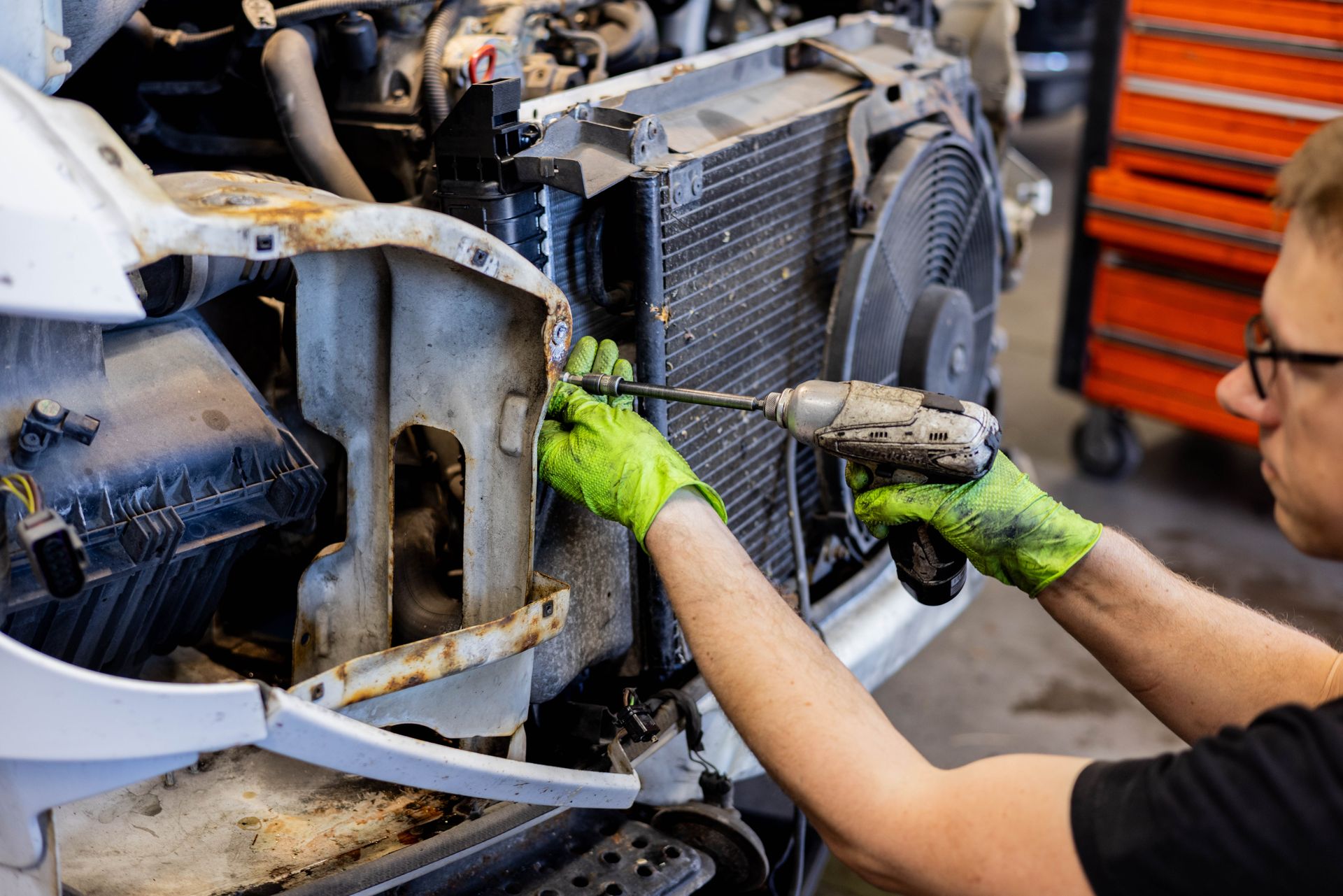 Mechanic with green gloves uses a power drill to work on the front of a vehicle in a repair shop. | Peak Euro