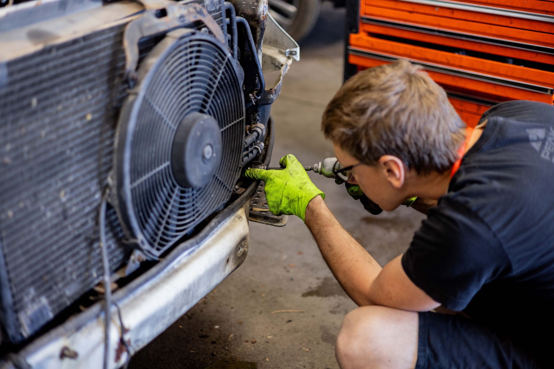Mechanic working on a car's radiator, using a drill. Wearing green gloves, in a garage setting. | Peak Euro