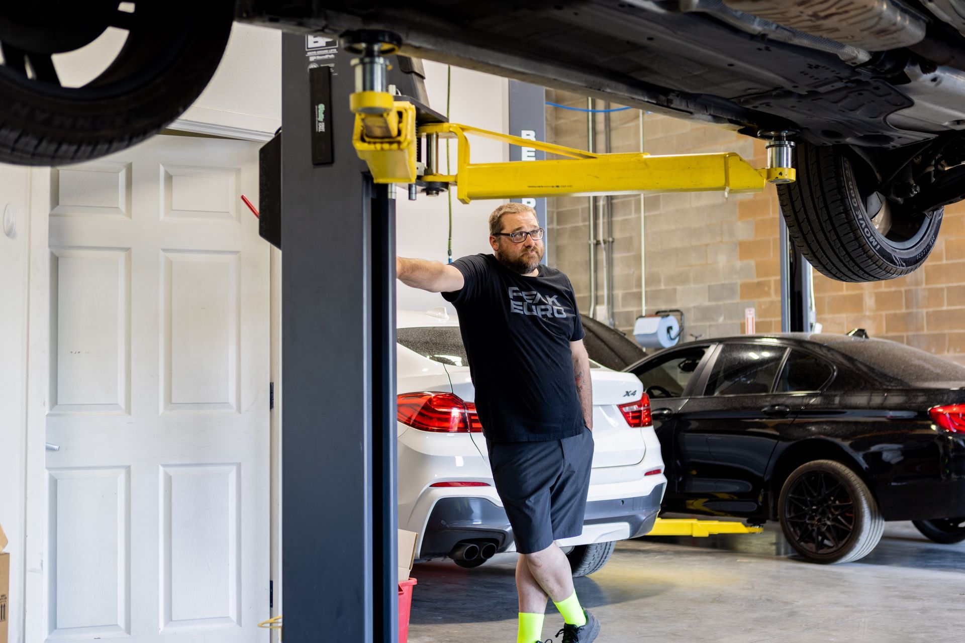Man in shop leans on car lift, under a vehicle; two cars and a door in background. | Peak Euro