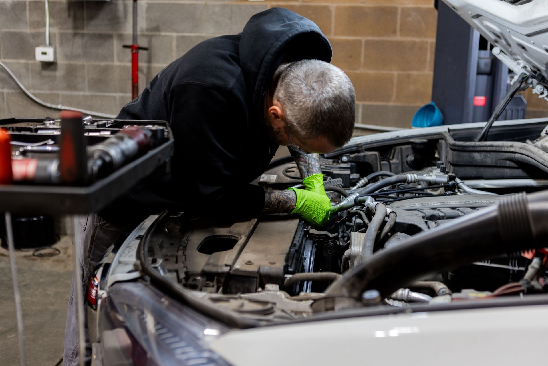 Mechanic in hooded jacket, wearing green gloves, working on a car engine in a garage.