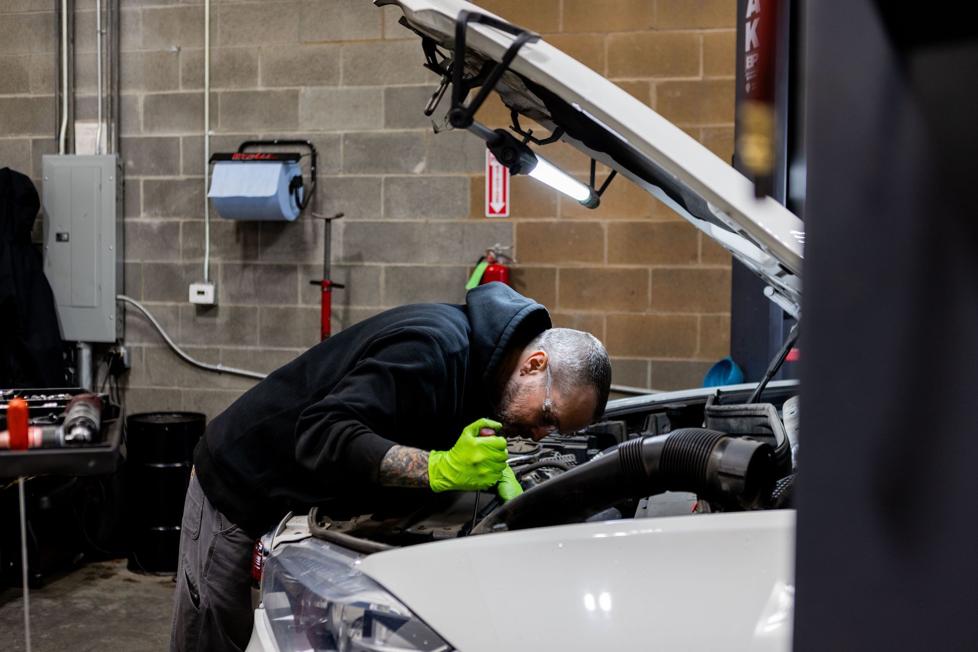 Mechanic working on a car engine in a garage, wearing a hoodie and gloves.