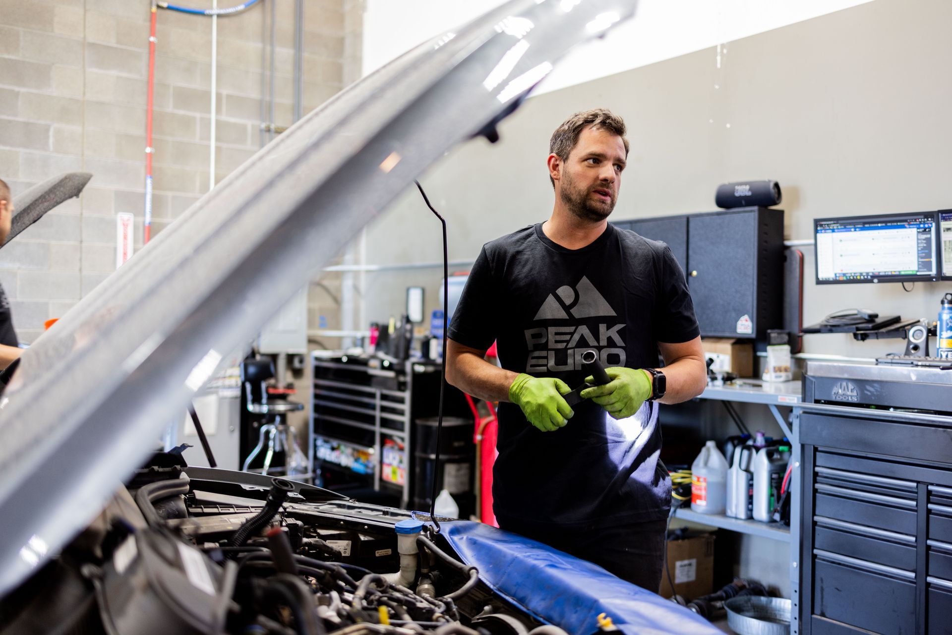Man in garage, car hood open, looking at engine. Wearing gloves, black t-shirt with mountain logo. | Peak Euro