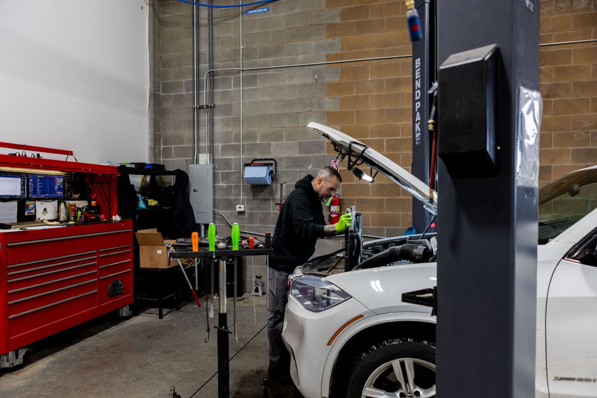 Mechanic working on a white car in a garage. He's wearing a black jacket and gloves. Tools are on a table.