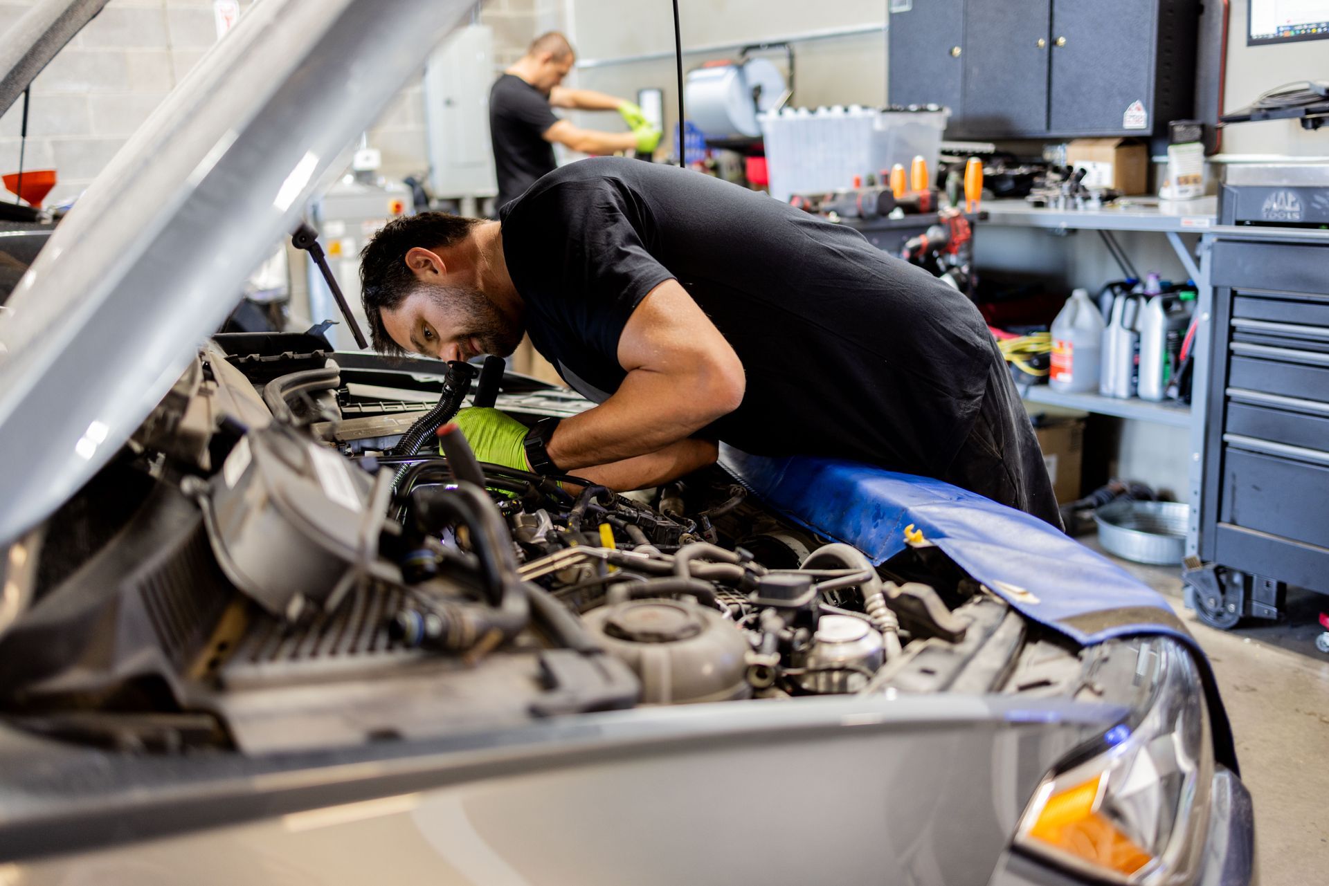 Mechanic working on a car engine in a garage, another person visible in the background. | Peak Euro