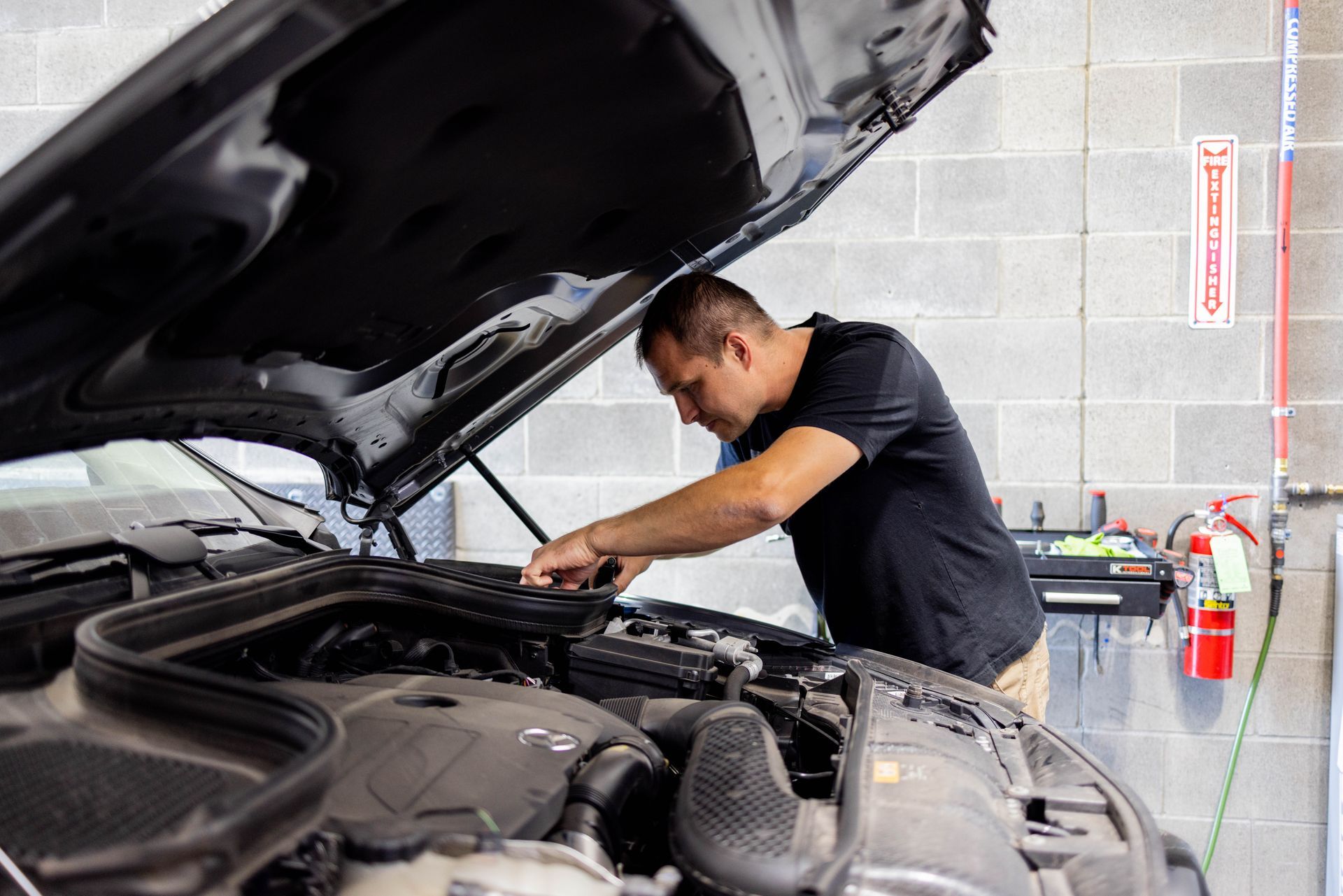 Mechanic working on a car engine, indoors; white brick wall, fire extinguisher, car hood open. | Peak Euro