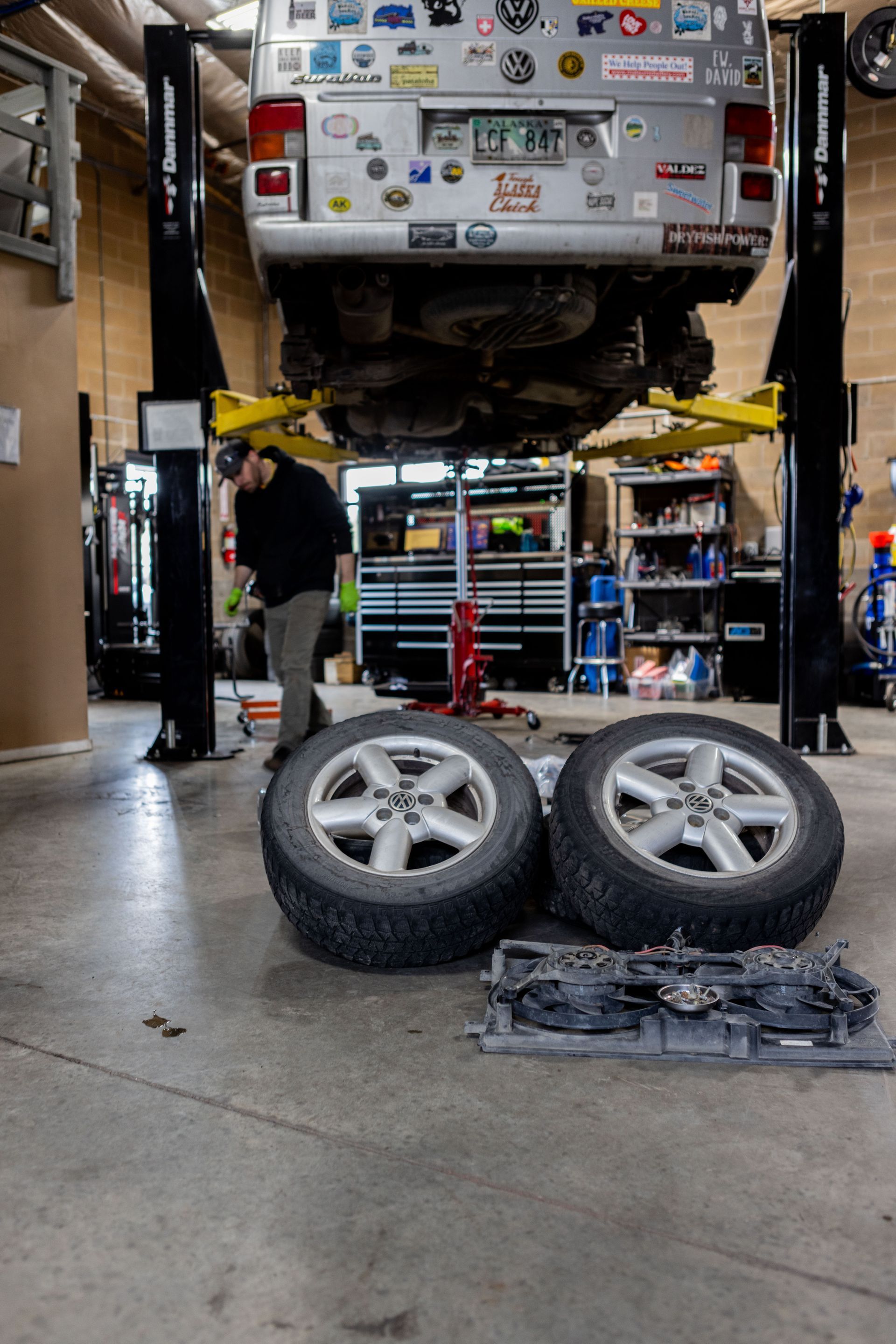 A van on a lift in a garage; a mechanic works on it. Two tires and a part sit on the floor. | Peak Euro