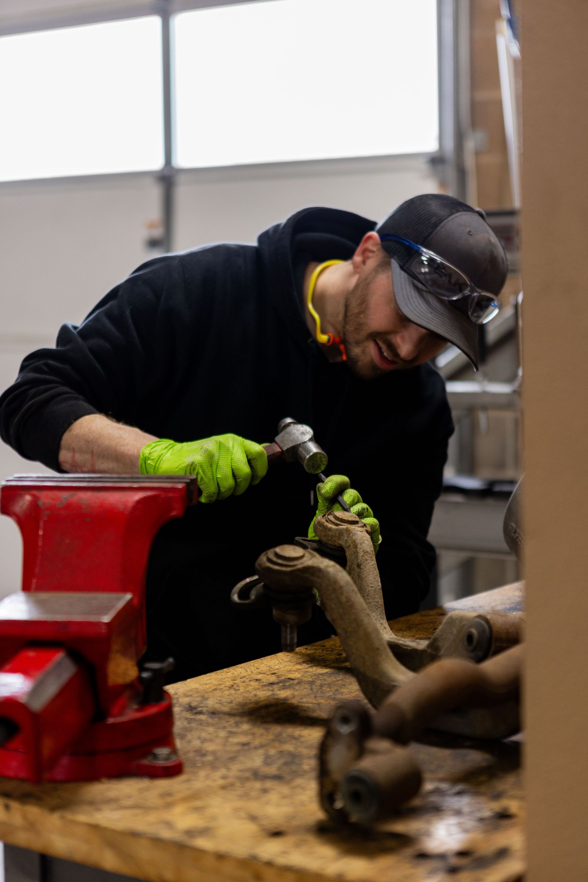 Man in safety glasses, cap, and gloves hammers on metal at a workbench, smiling. | Peak Euro