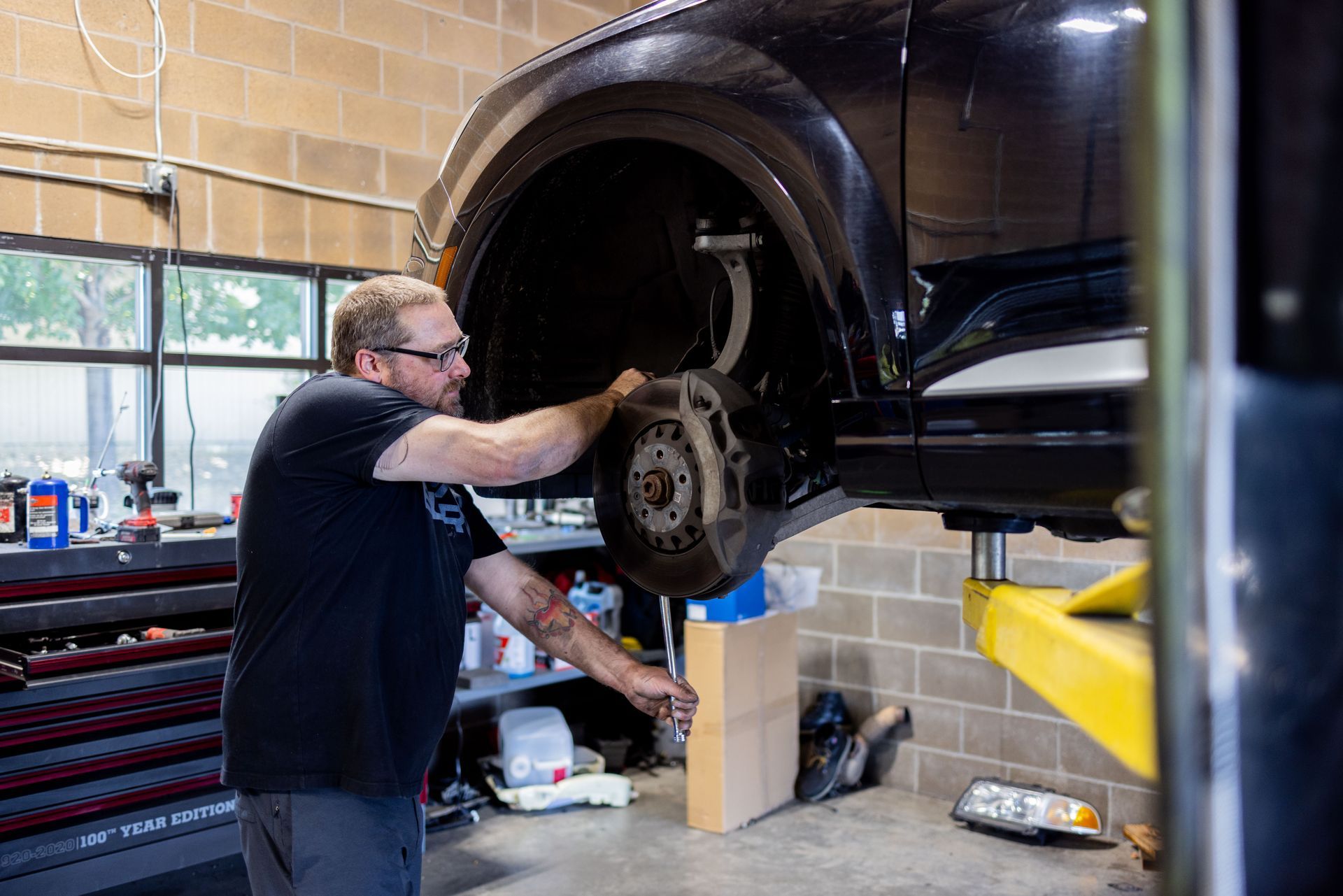 Mechanic working on a car's brakes in a garage. He wears glasses and a black shirt. The car is lifted.