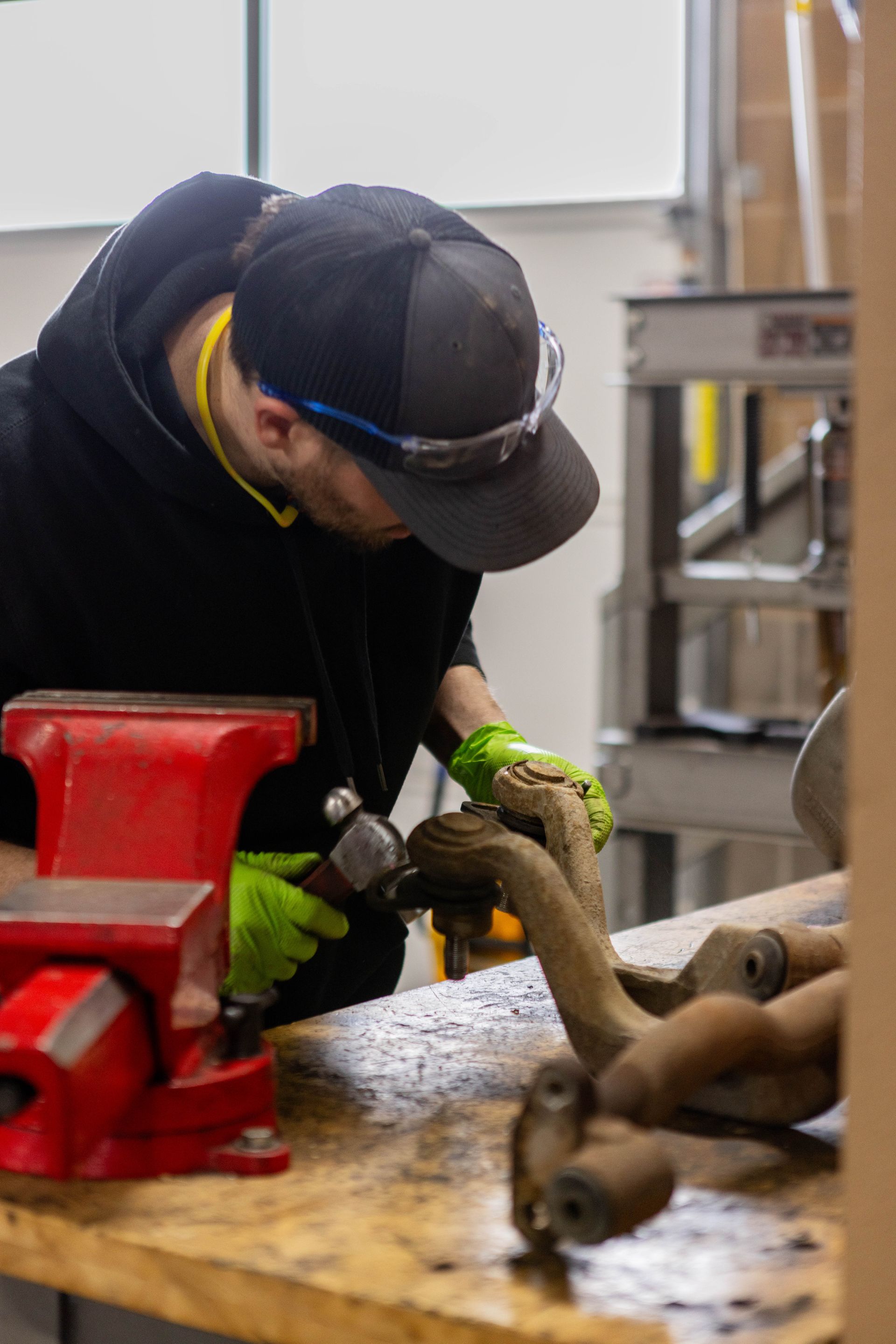 Person in black hoodie and hat, wearing safety glasses and gloves, working on metal piece with hammer at a workbench. | Peak Euro