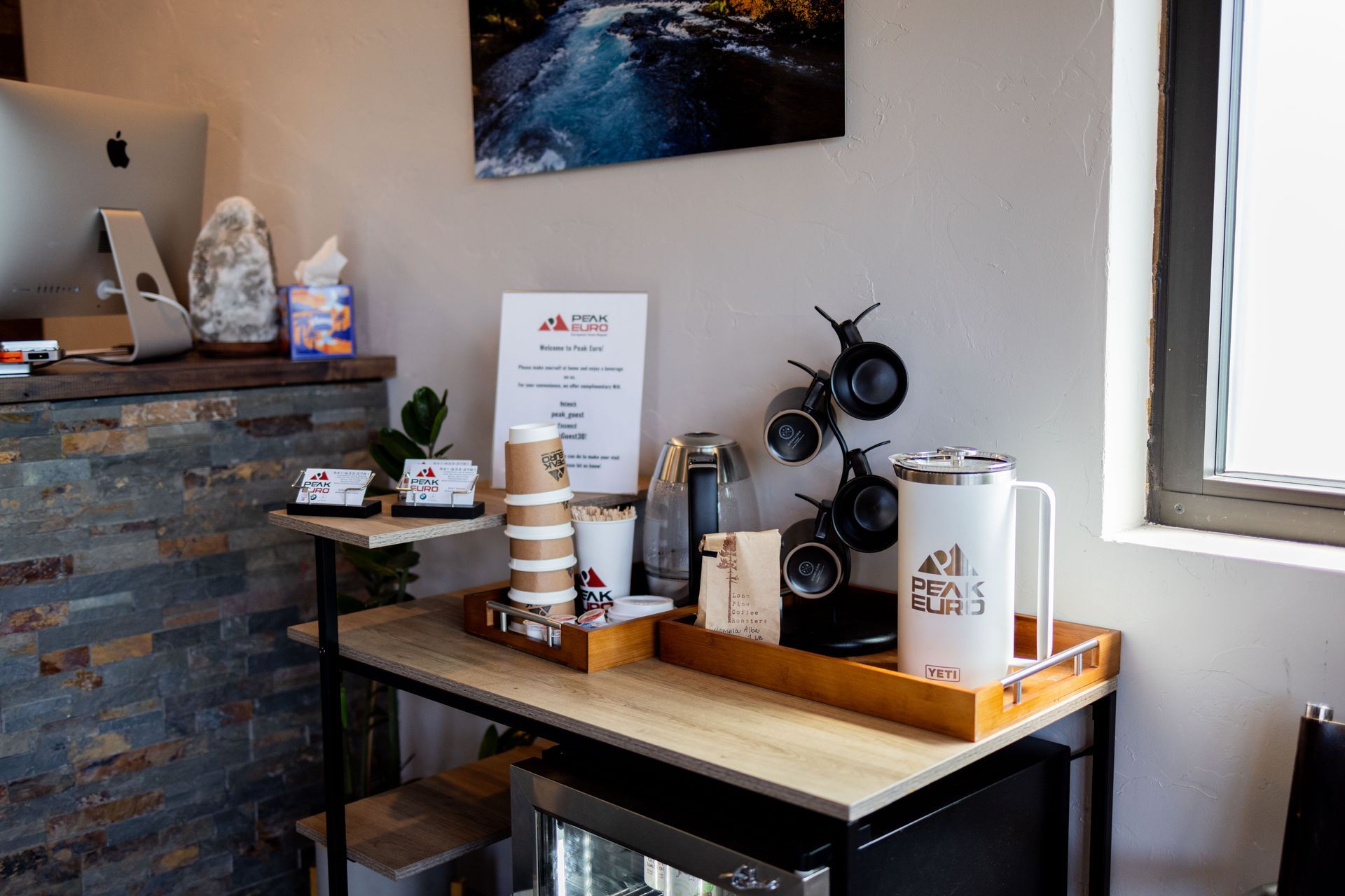 Coffee station: desk with coffee cups, a dispenser, a thermal, a mini-fridge, and a painting. | Peak Euro