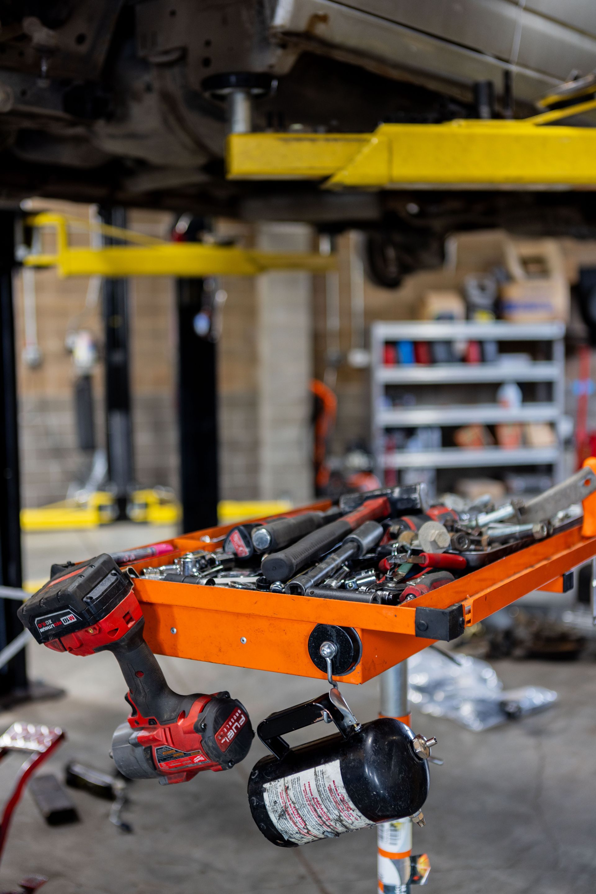 Orange tool cart with tools, a red drill, and an oil can in an auto repair shop; a car is raised on a lift. | Peak Euro