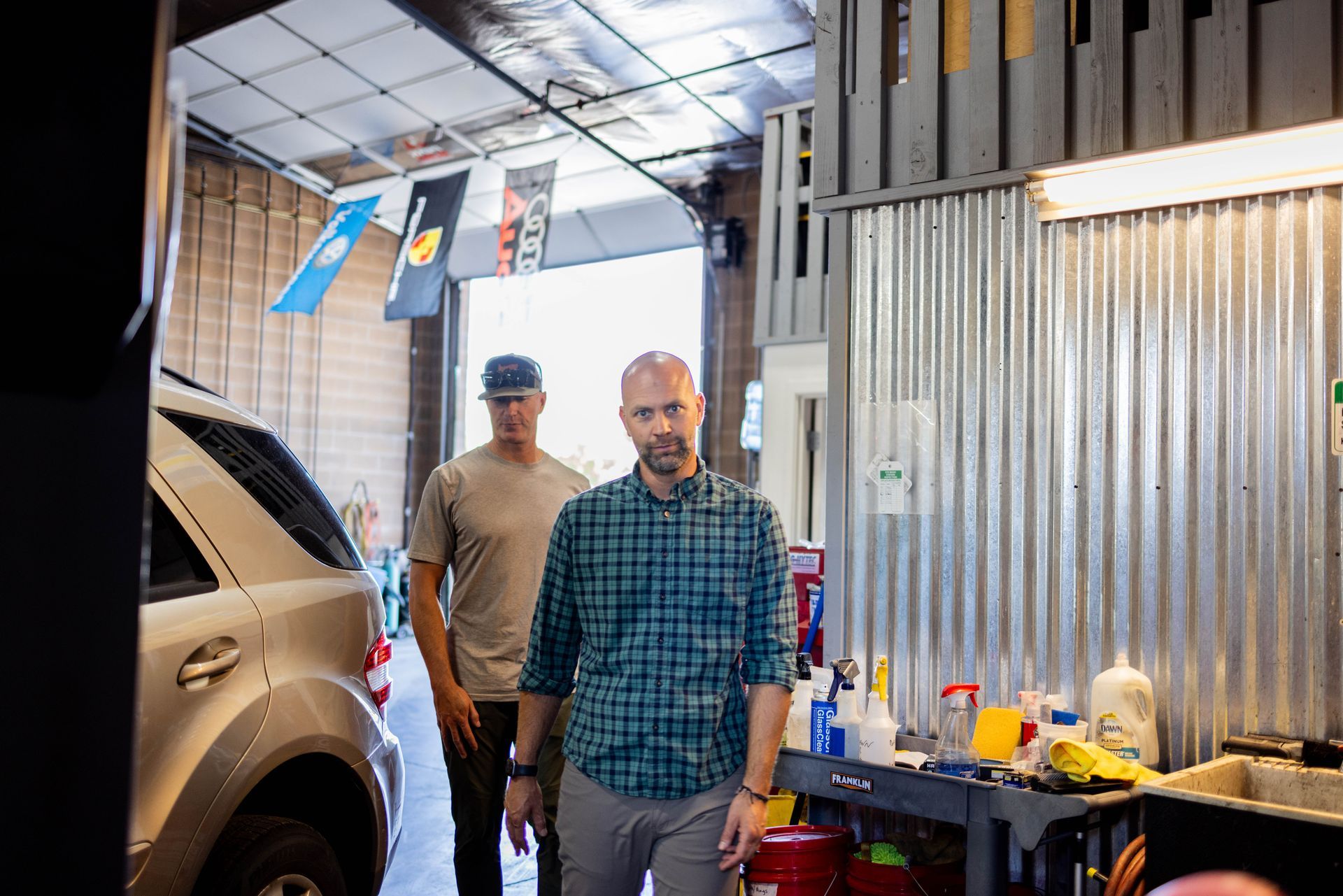 Two men in a garage. One in a plaid shirt walks toward the camera, the other in a hat follows. | Peak Euro