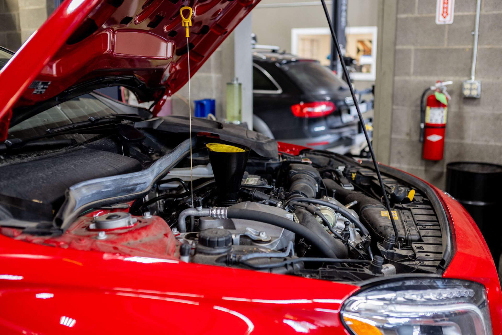 Red car with hood open in a garage, engine visible. Another car and fire extinguisher in the background. | Peak Euro