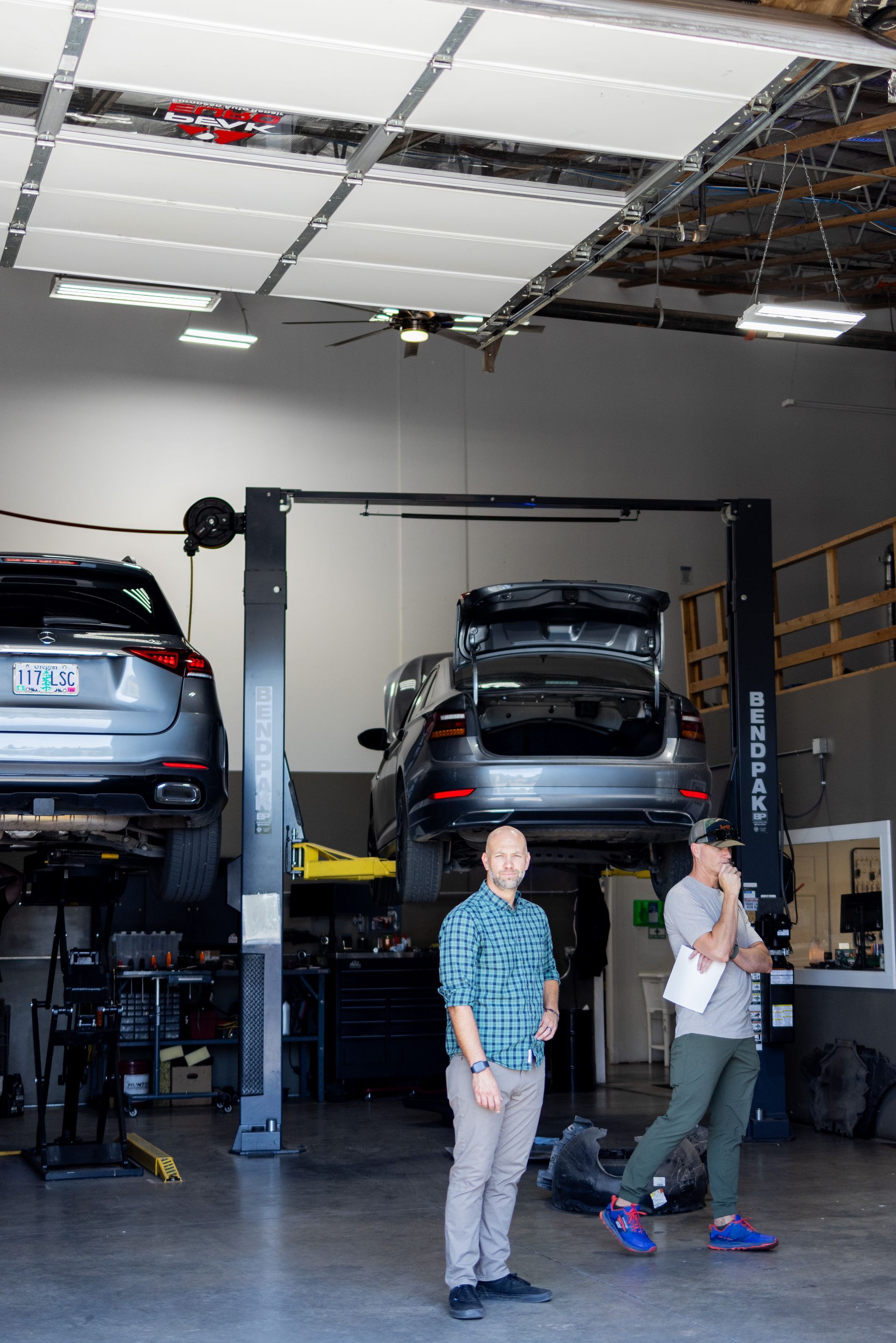 Two men in a garage with cars on lifts. One man smiles in a blue plaid shirt; the other holds notes. | Peak Euro