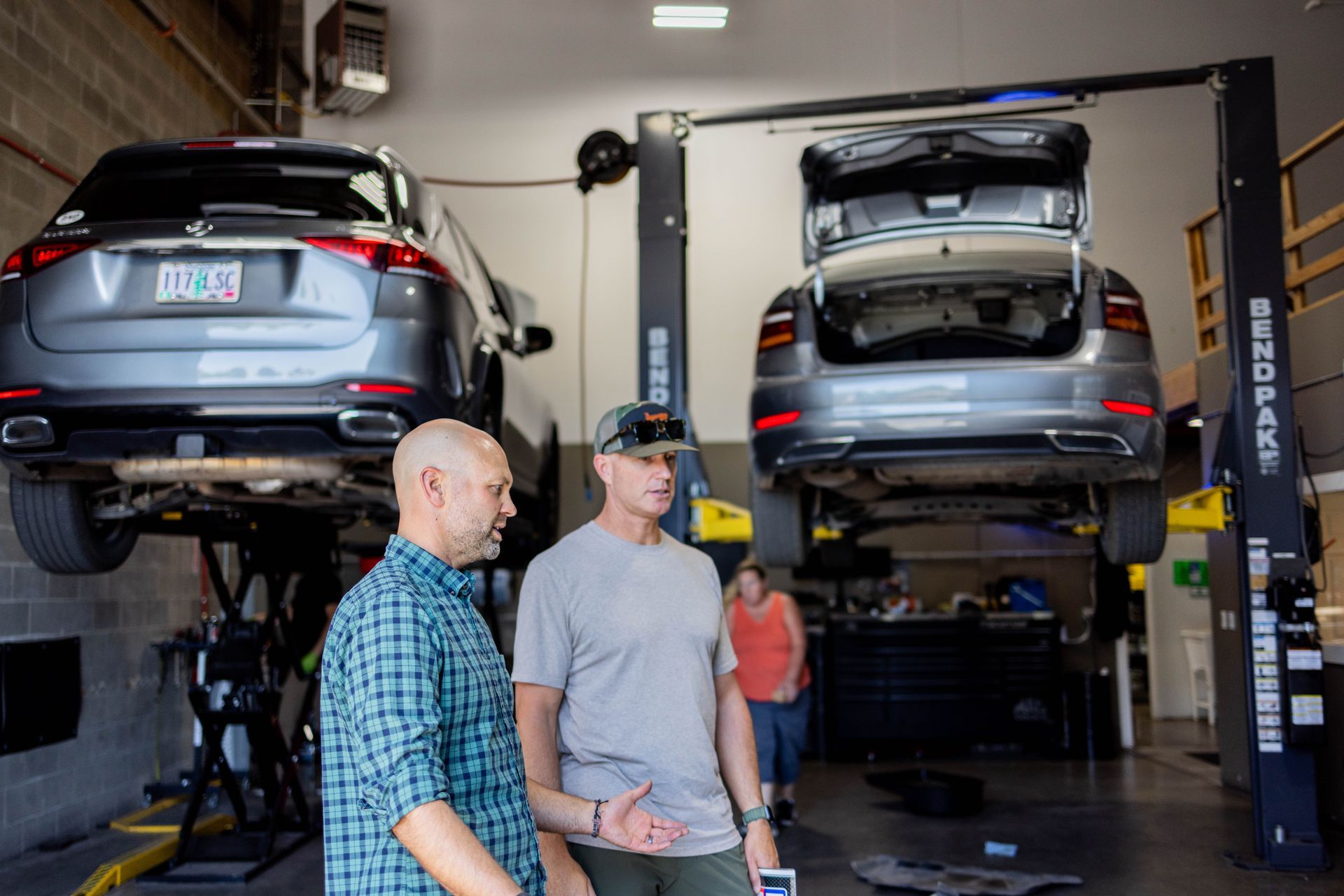 Two men in a garage, looking at cars on lifts. One man gestures, discussing the vehicles' mechanics. | Peak Euro