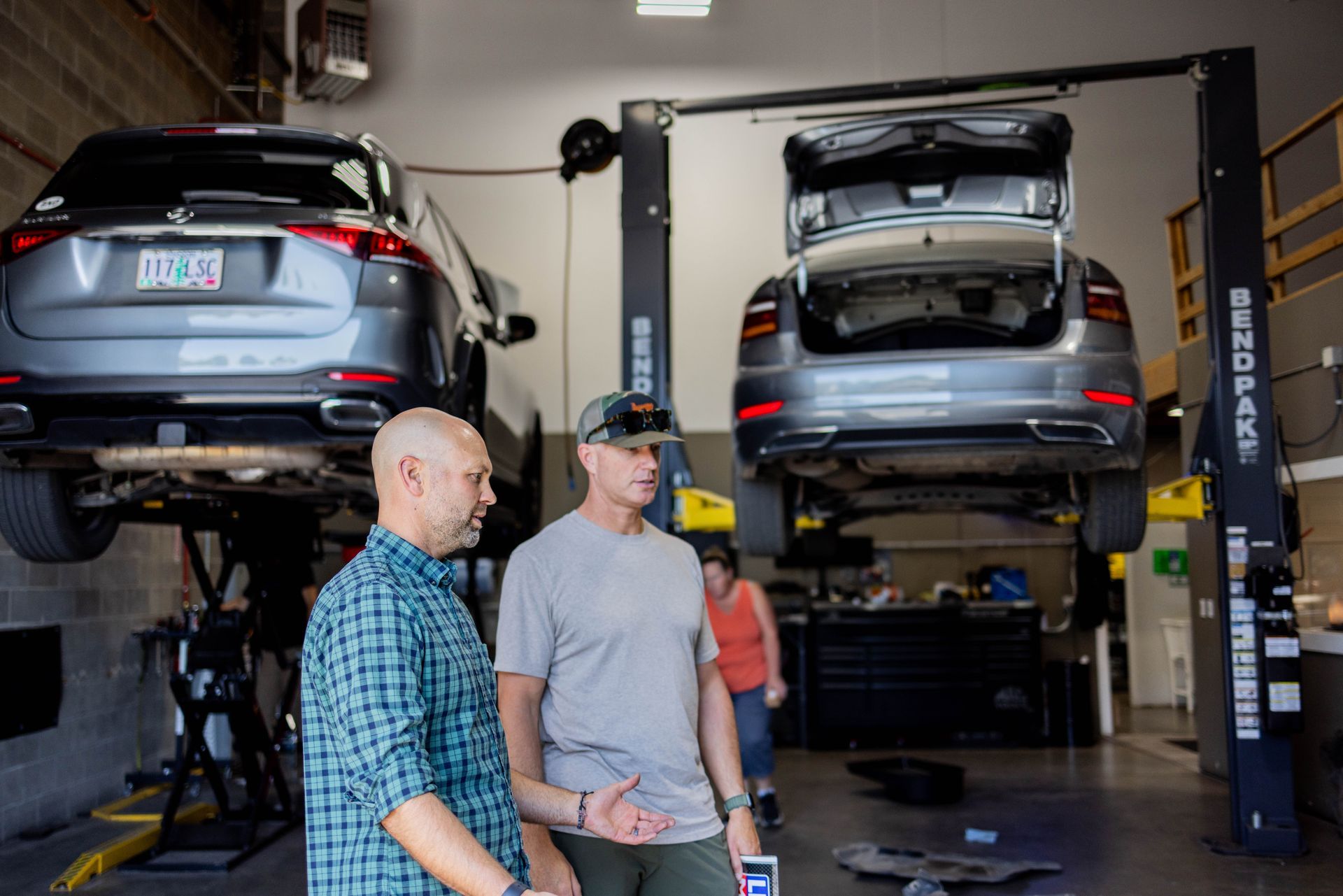 Two men in a car repair shop, looking at a car on a lift. Another person walks in the background. | Peak Euro