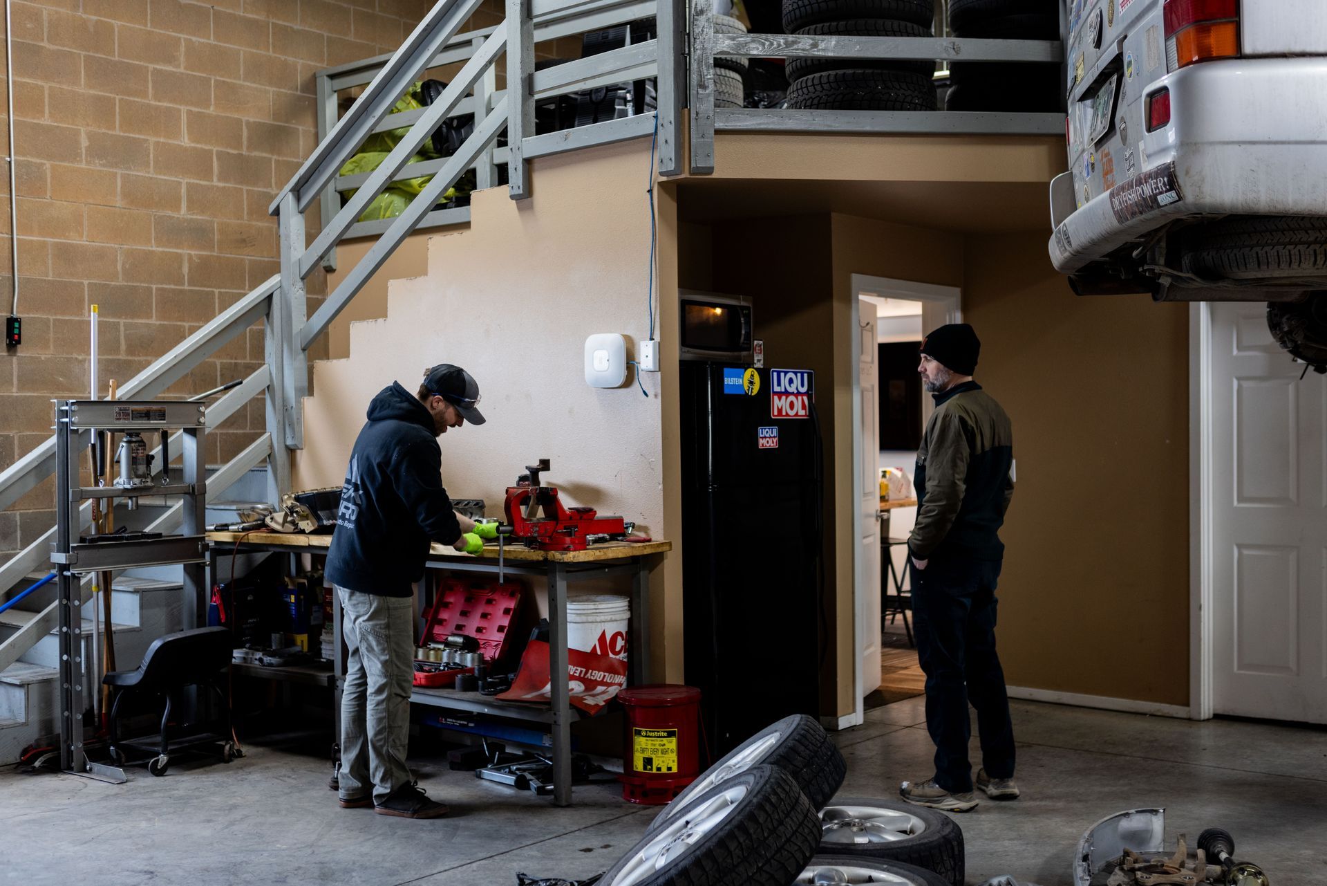 Two men in a garage, one working at a workbench, the other standing. Tools and car parts visible. | Peak Euro