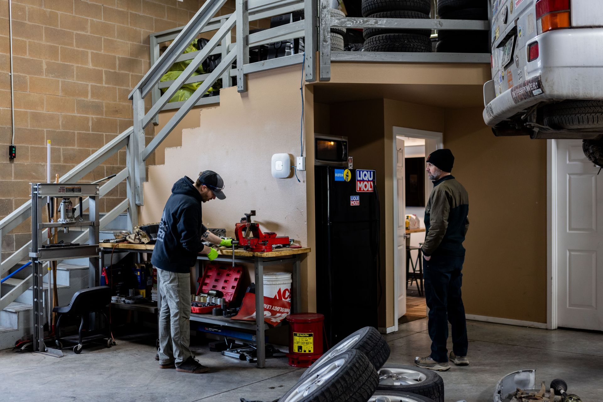 Two men in a garage working on a vehicle. One at a workbench, the other standing. A second-story storage area is above.| Peak Euro