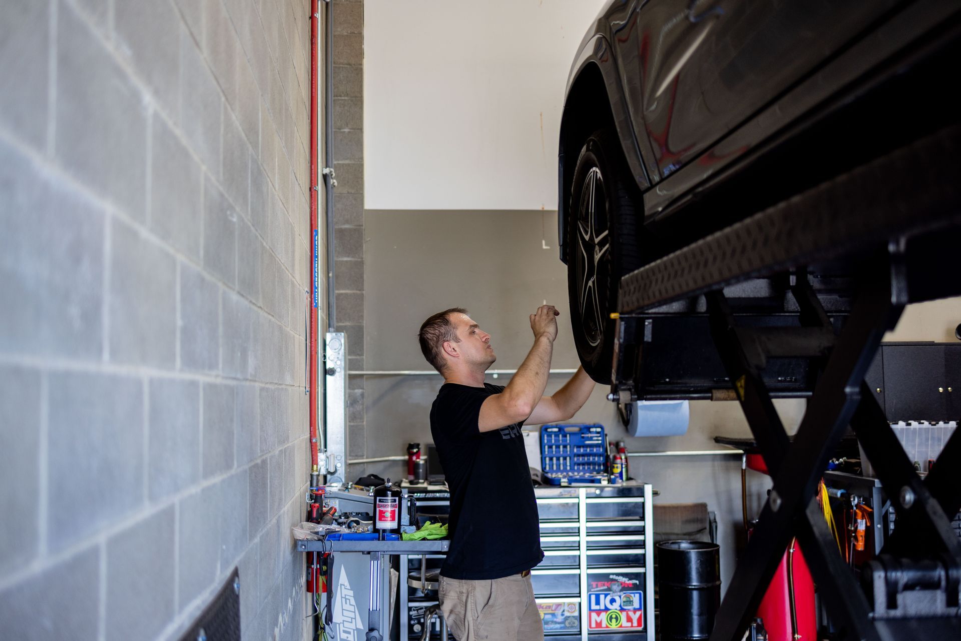 Mechanic working on a car lifted in a garage. Wearing black shirt and khaki pants. Concrete block walls. | Peak Euro