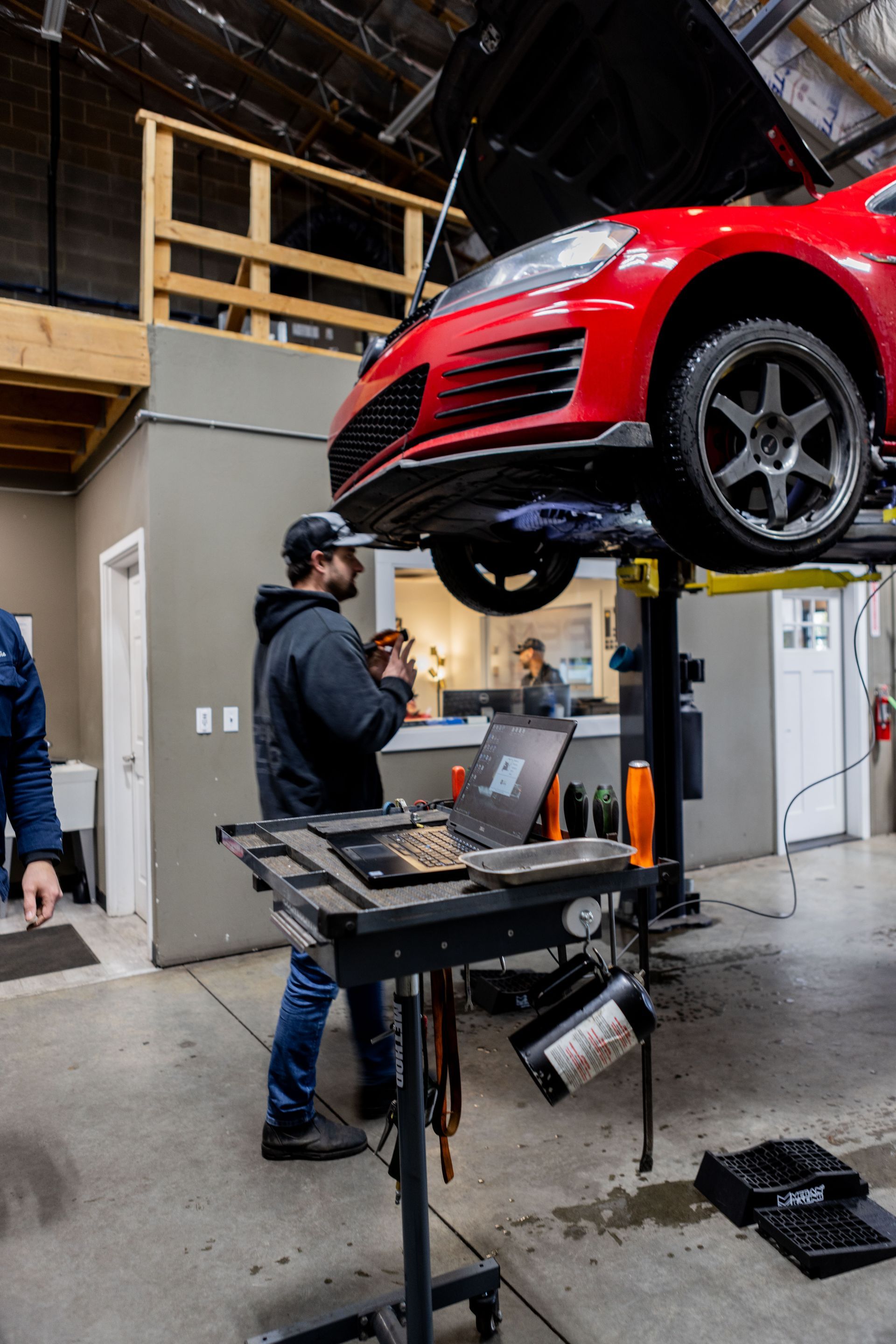 Mechanic working on a red car lifted in a garage. He is using a laptop on a rolling cart.| Peak Euro