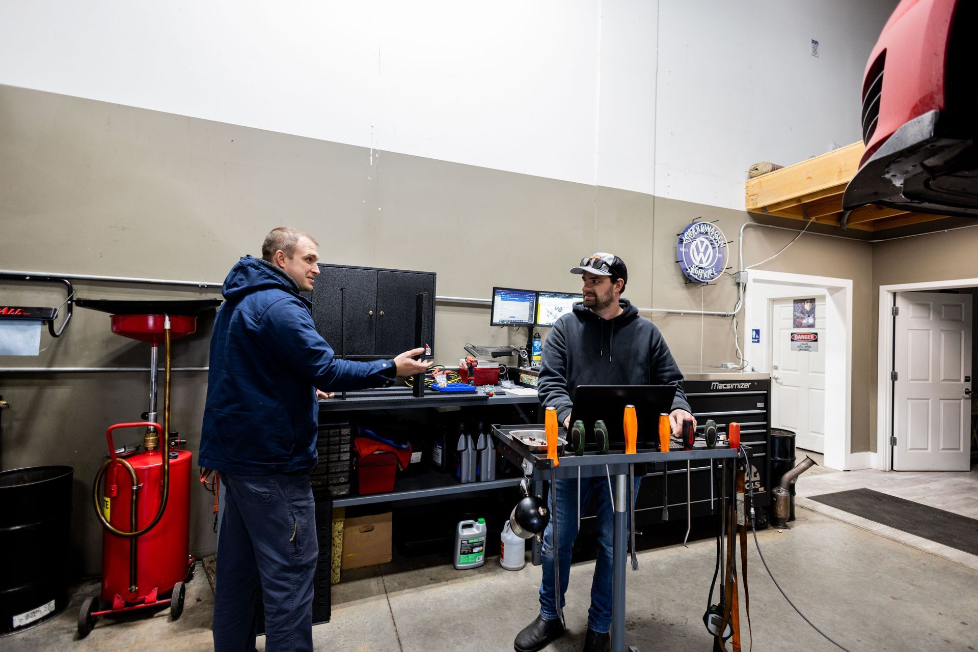 Two mechanics in a garage discussing by a workstation. One points at a monitor. Car is raised overhead.