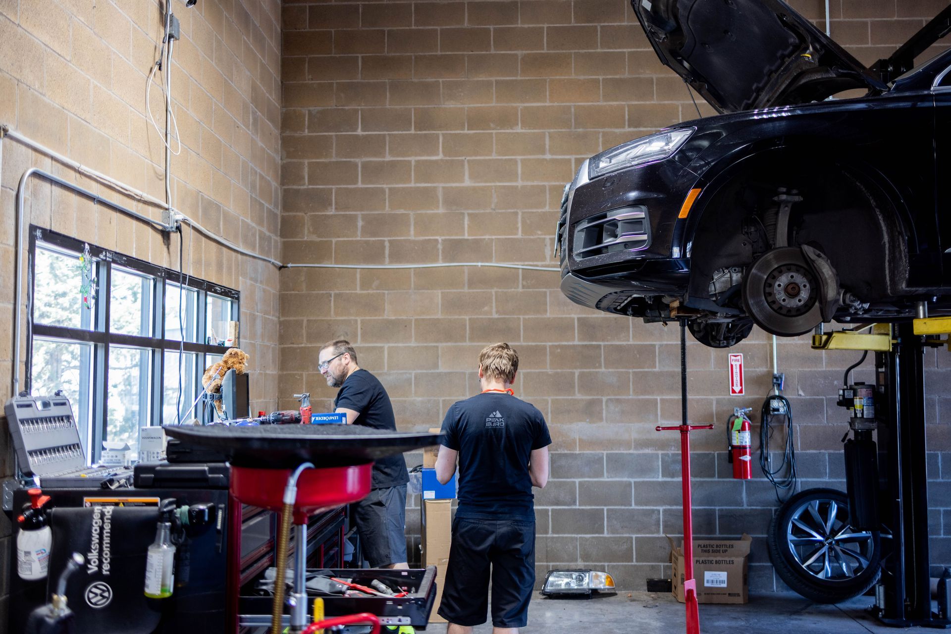Two mechanics working on a car in a garage; car on a lift, tools, brick wall background. | Peak Euro