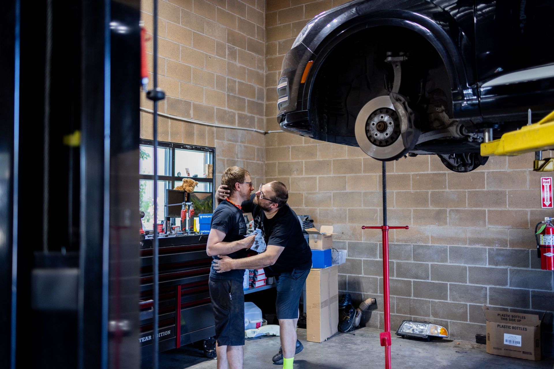 Two men embrace and kiss in an auto repair shop with a car on a lift. | Peak Euro