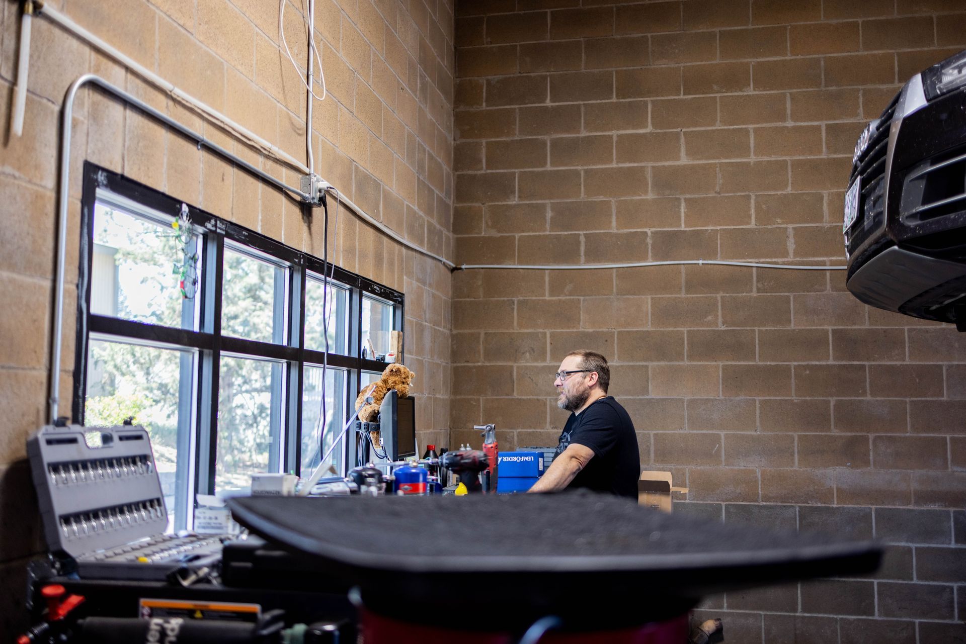 Man in glasses at a desk in a garage, near a window and a car lift. Brick walls surround. | Peak Euro