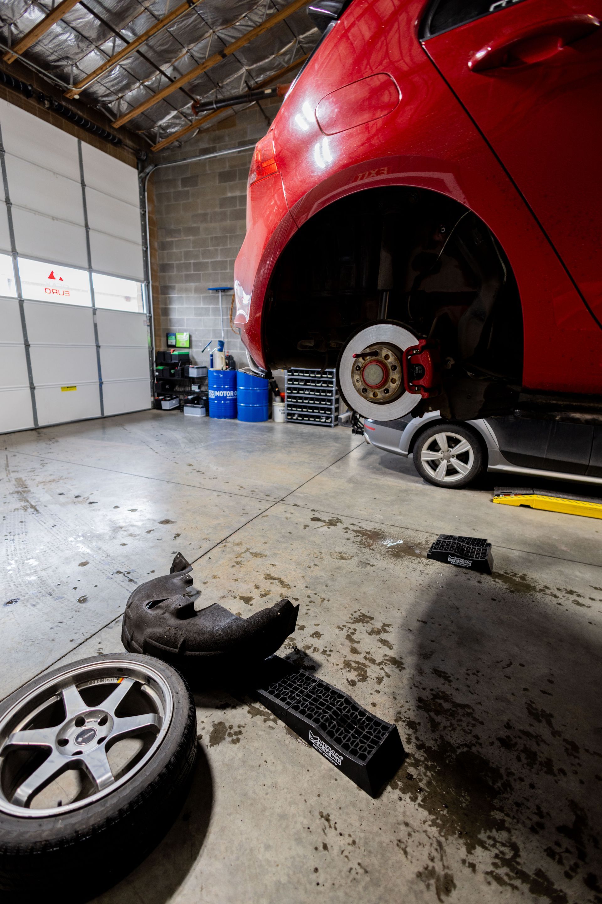 Red car being repaired in a garage; a wheel and parts lie on the floor.| Peak Euro