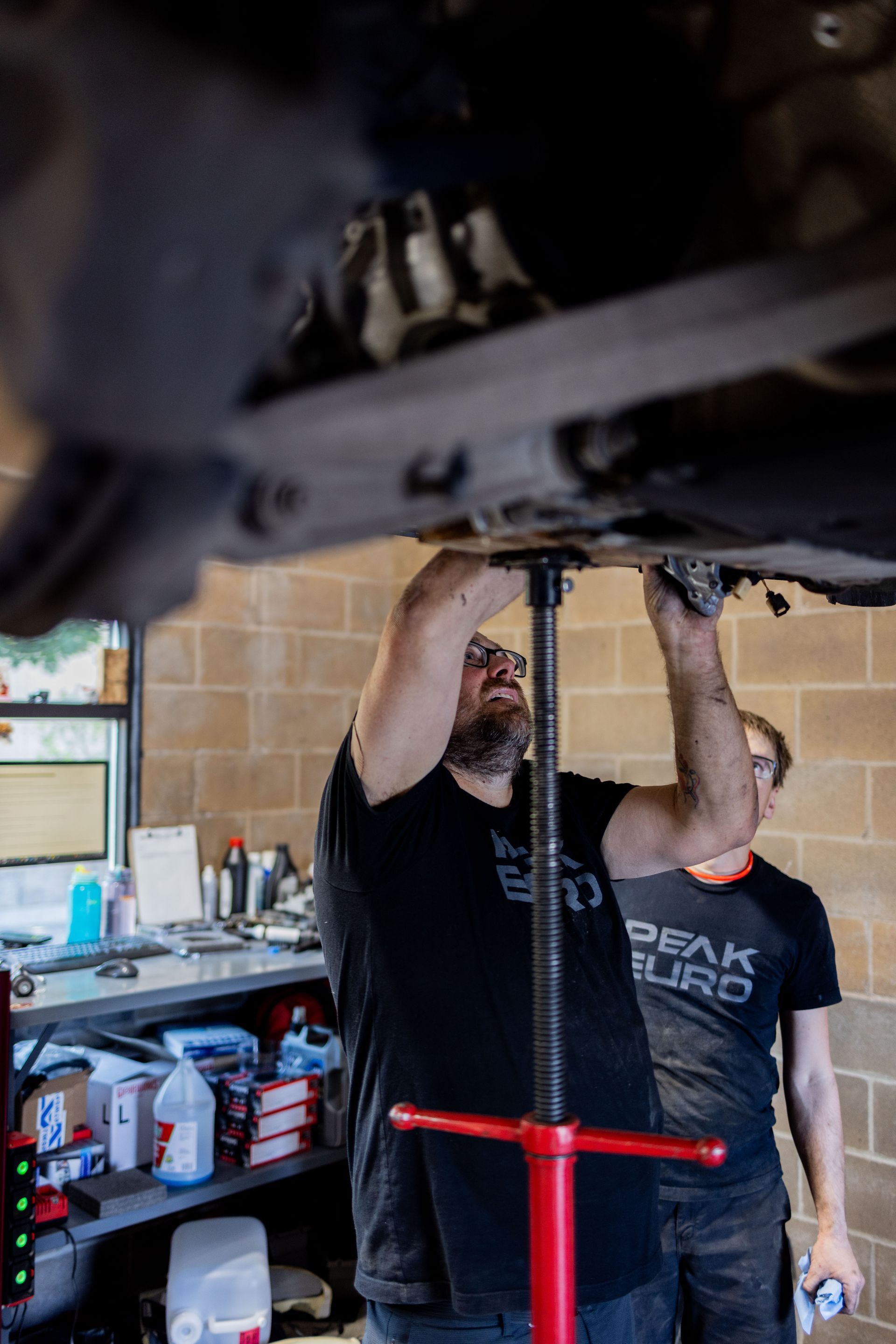 Man in black shirt working under a car, using a jack. Another man watches. Garage setting. | Peak Euro