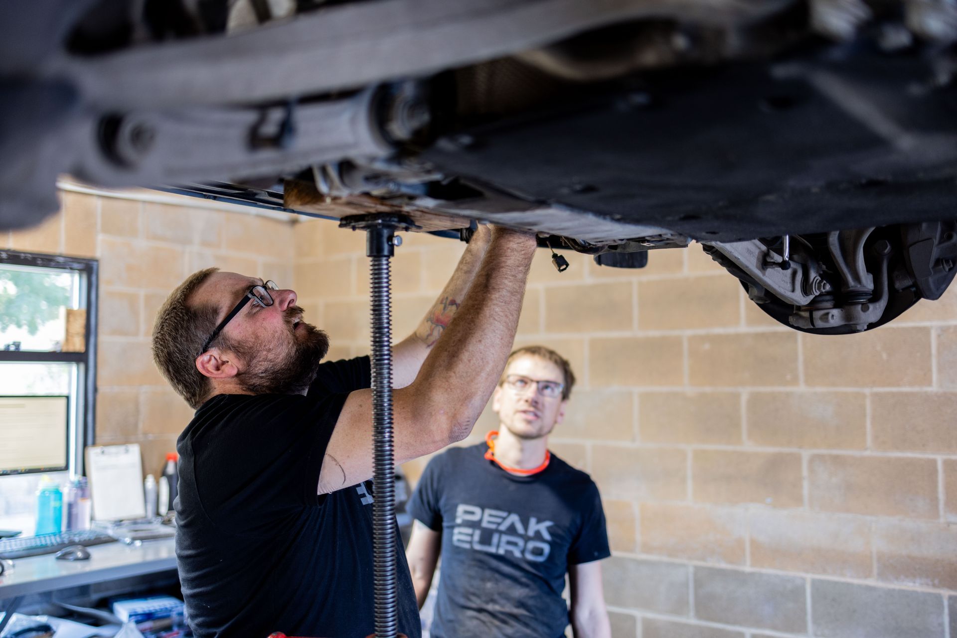 Two men working under a car; one is using a jack. Workshop setting. | Peak Euro