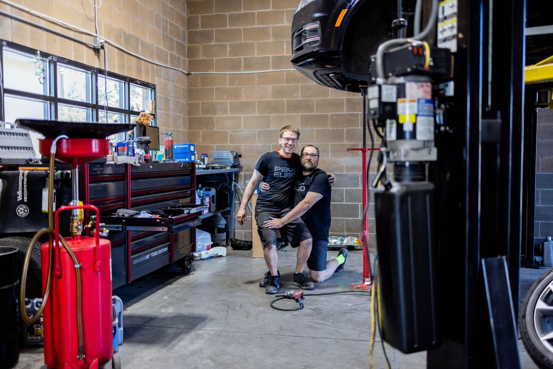 Two men in a garage posing by a car on a lift; one kneeling, one standing, smiling. | Peak Euro