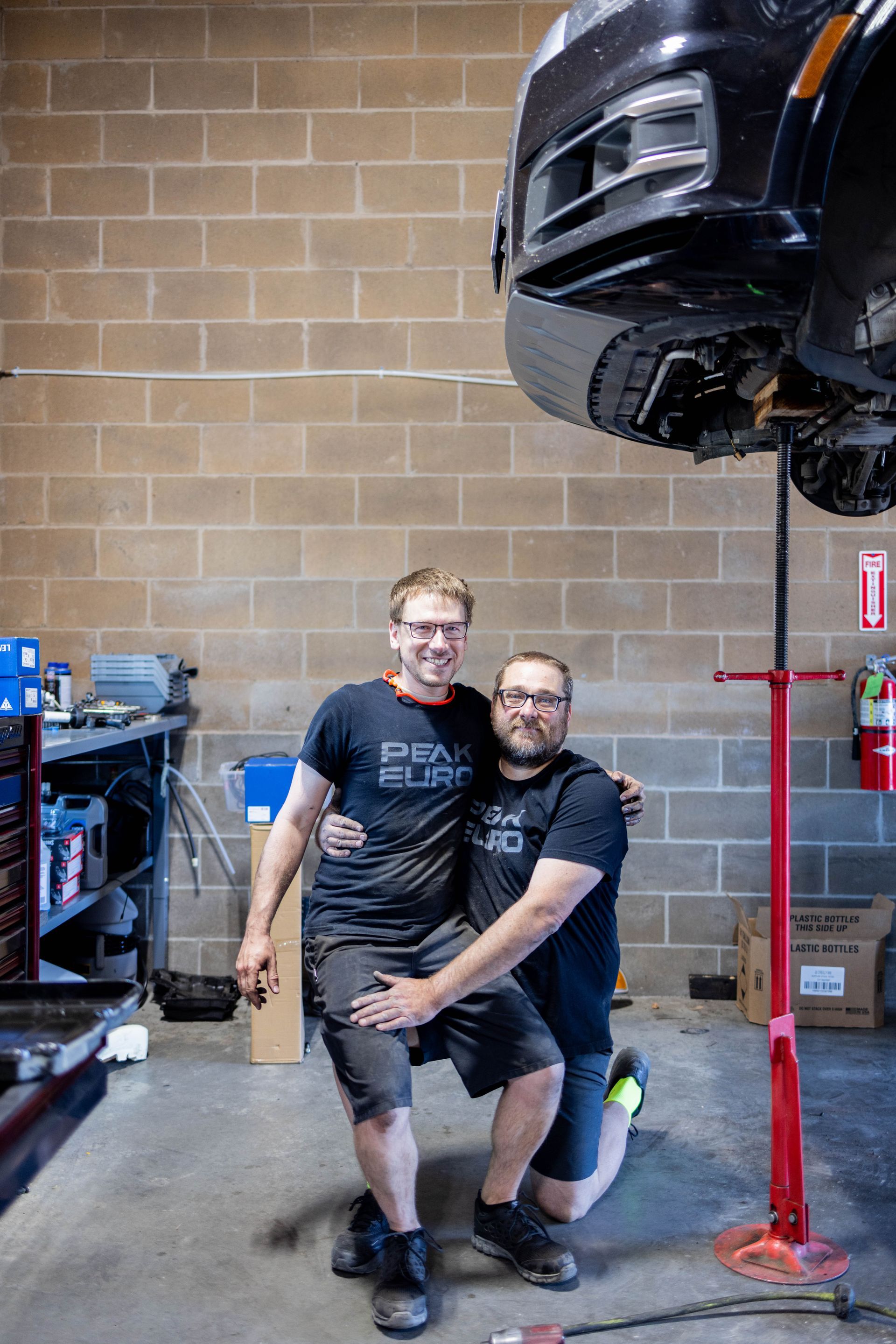 Two men pose in auto shop. One kneels, hugging the other's waist under a lifted car. | Peak Euro