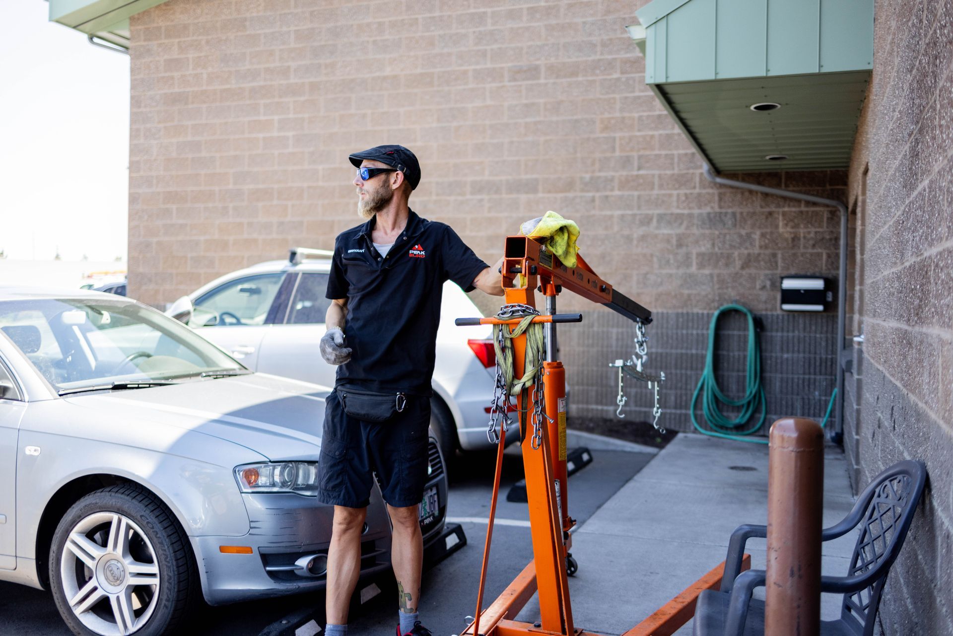 Man in black shirt and shorts standing near an orange engine hoist next to a car outside a building. | Peak Euro