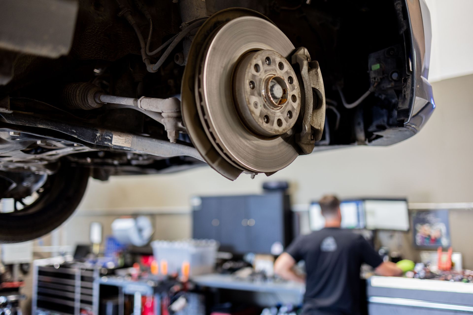 Car's brake rotor and caliper exposed, mechanic working in background in a garage setting. | Peak Euro