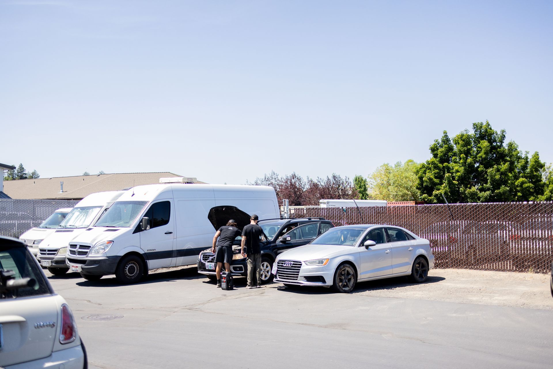 Two men examining a car next to a white van and a silver car on a sunny day.