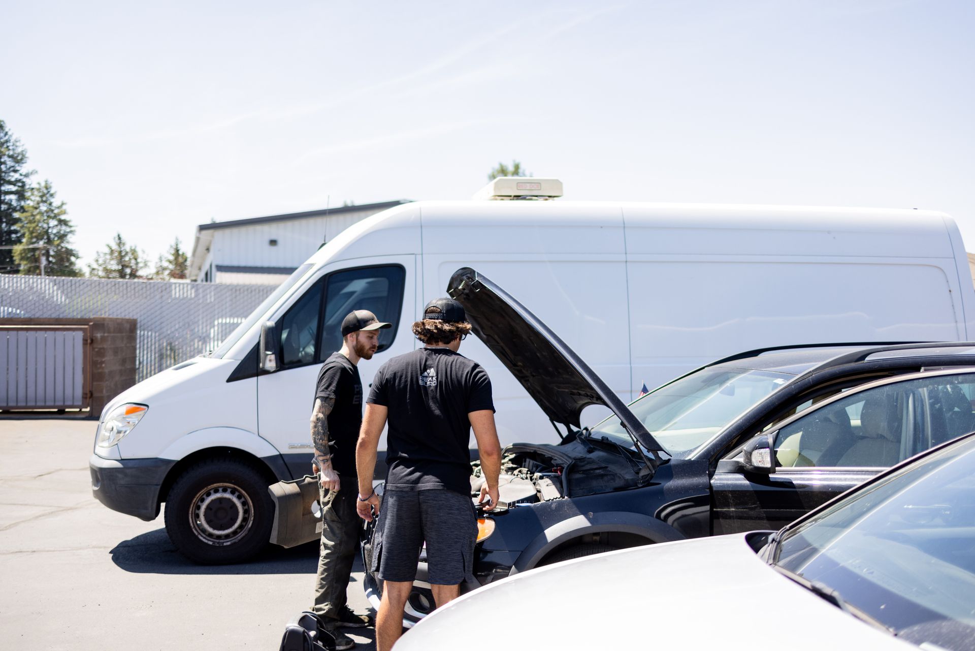 Two men examining a car engine with a white van in the background on a sunny day. | Peak Euro
