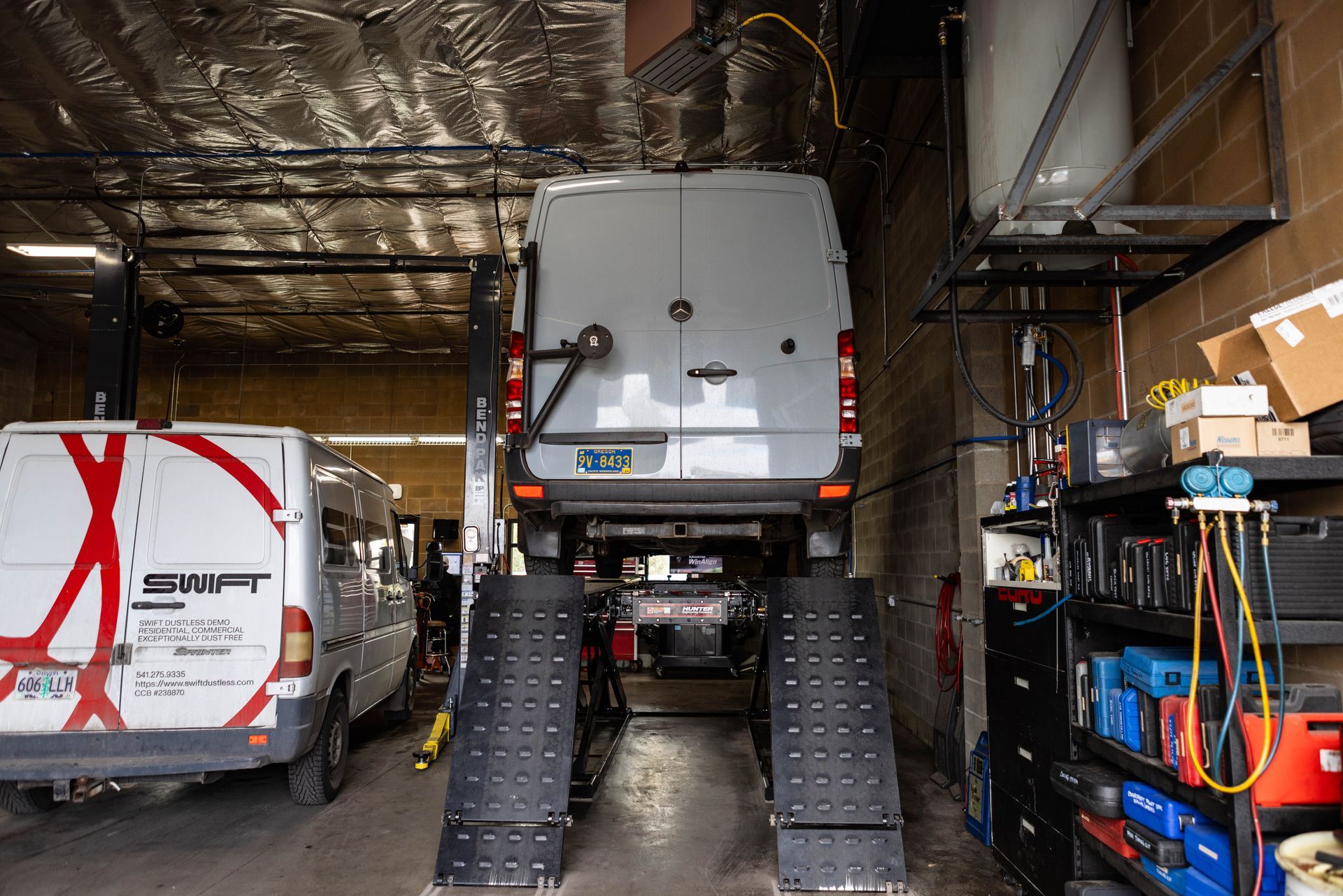 White van on a lift in a garage, next to another van. Tools and shelving are visible. | Peak Euro