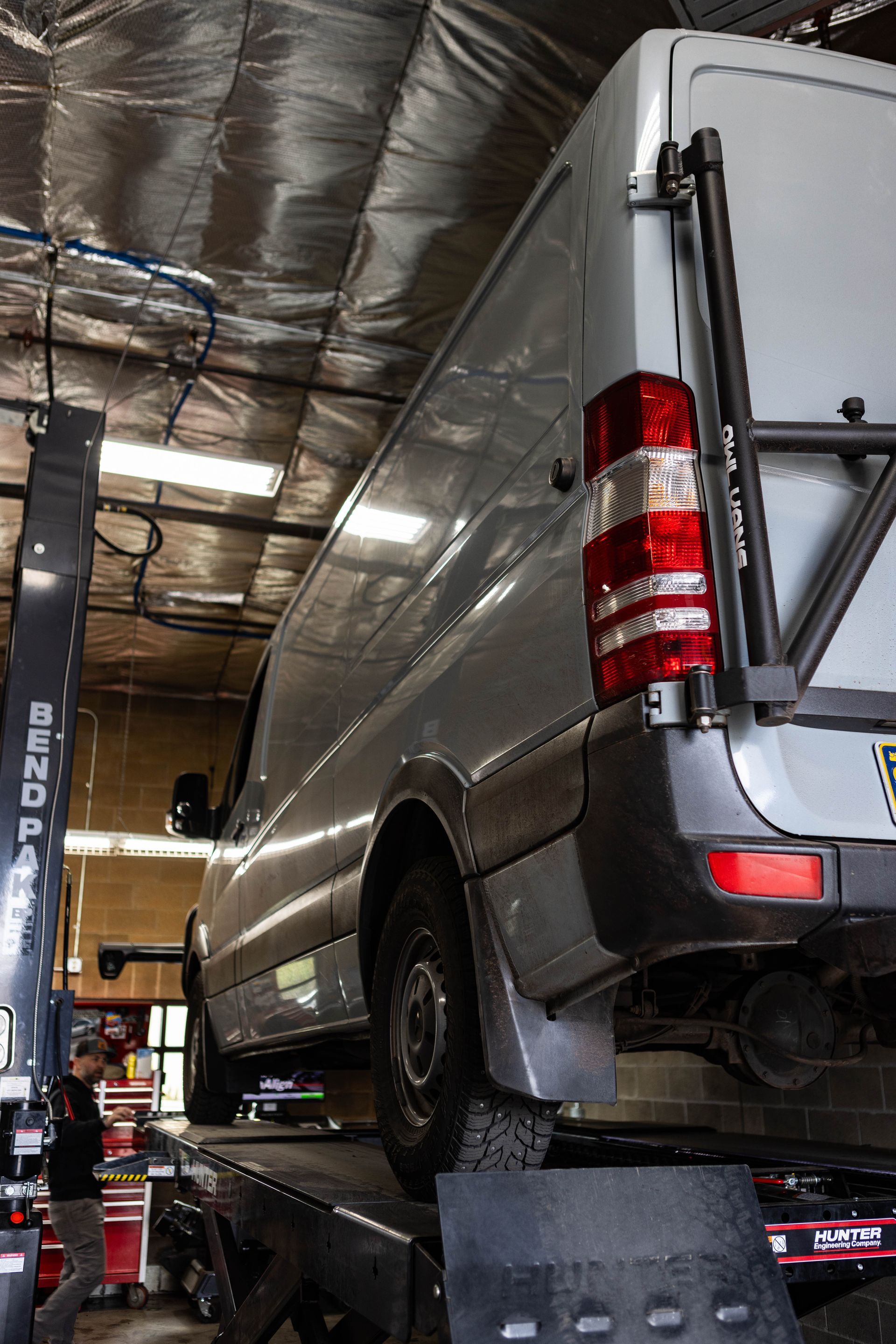 A silver van raised on a lift in a workshop, a person stands to the left, red tool chest in background. | Peak Euro