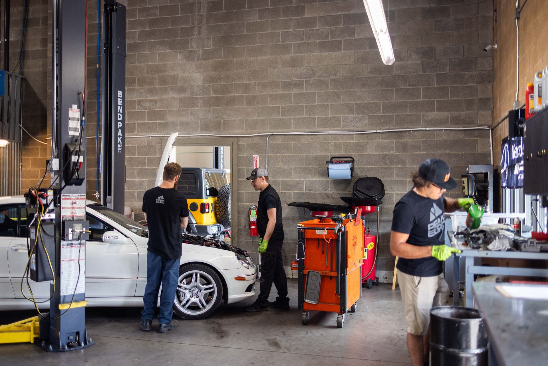 Three mechanics working on a white car inside an auto repair shop. One is under the hood, the others are near tools. | Peak Euro