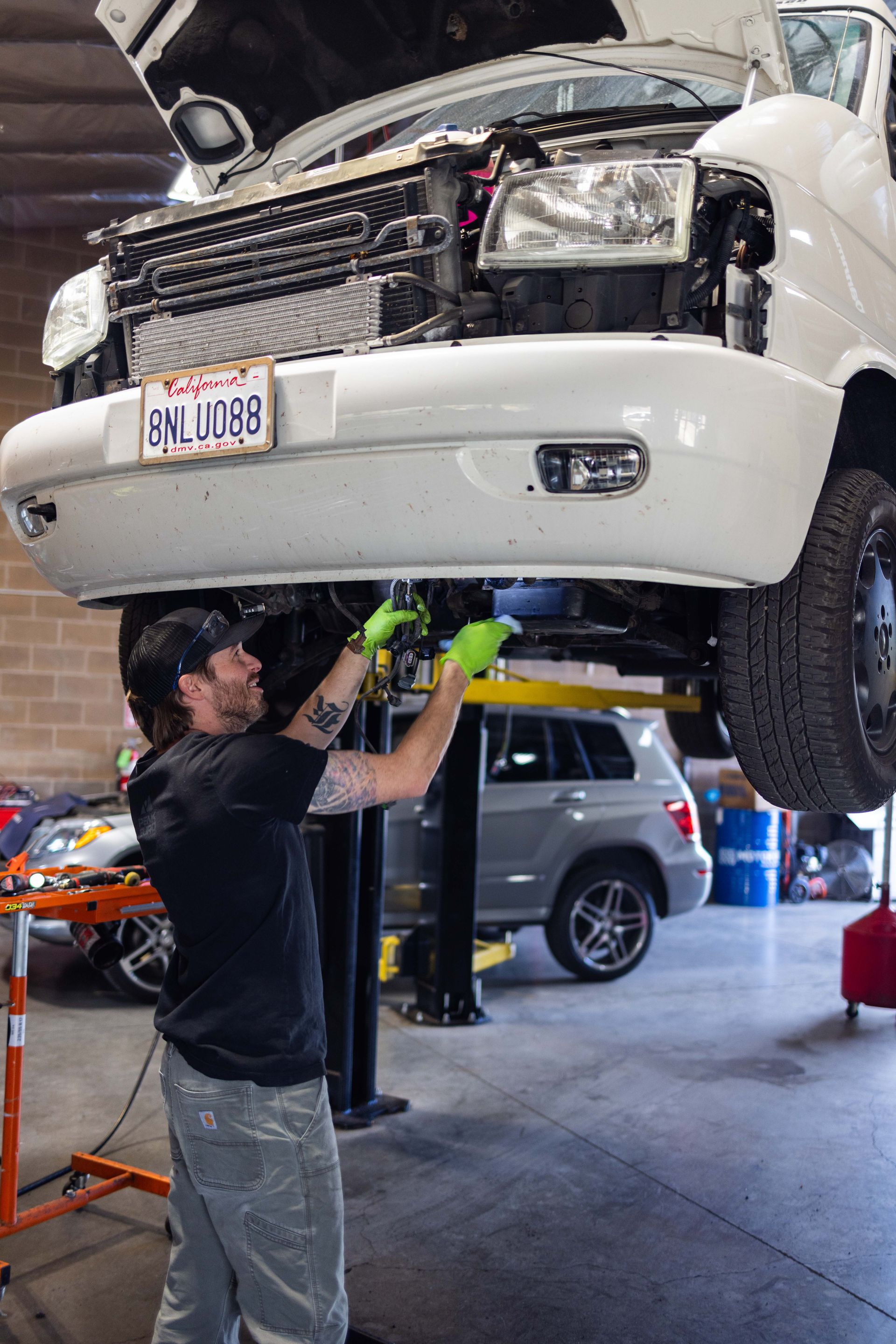 A mechanic in green gloves works on a white van lifted in a garage, smiling. | Peak Euro