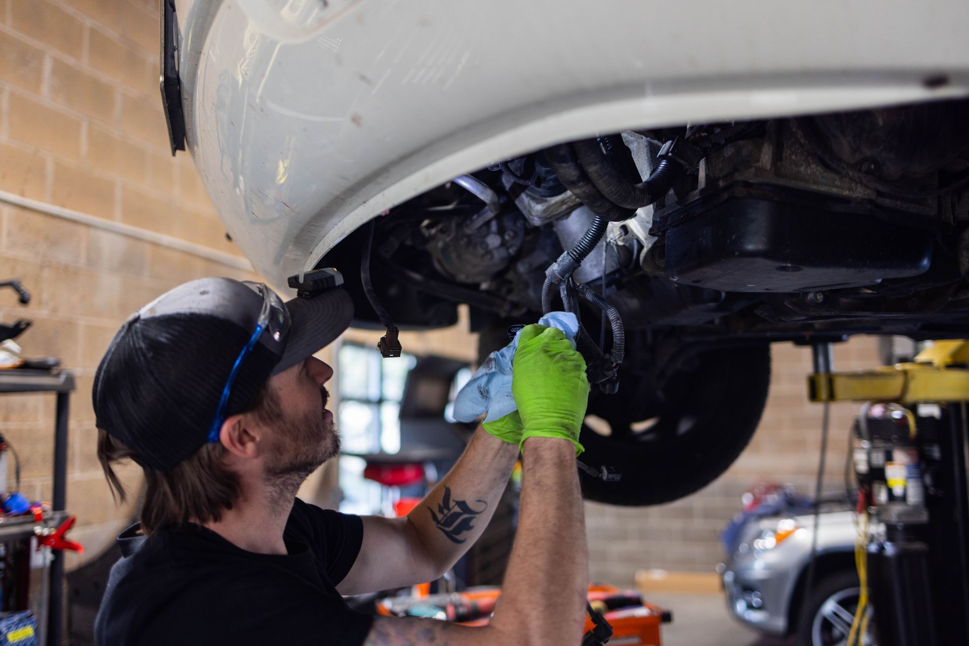 Mechanic in green gloves, working under a white car, wiping engine parts in a garage. | Peak Euro